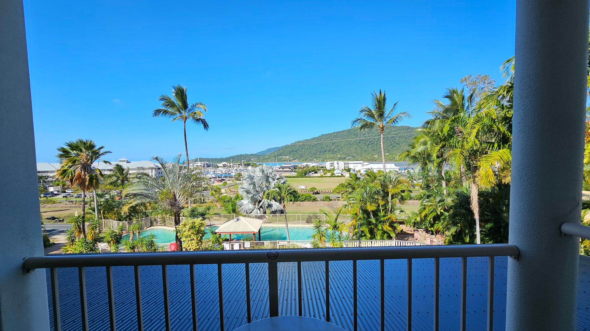 View from a balcony with a railing in the foreground. Below, a swimming pool is surrounded by palm trees and a small gazebo. In the middle distance, there are buildings and a marina with boats. A lush green hill rises in the background under a clear blue sky. View from a balcony with a railing in the foreground. Below, a swimming pool is surrounded by palm trees and a small gazebo. In the middle distance, there are buildings and a marina with boats. A lush green hill rises in the background under a clear blue sky.