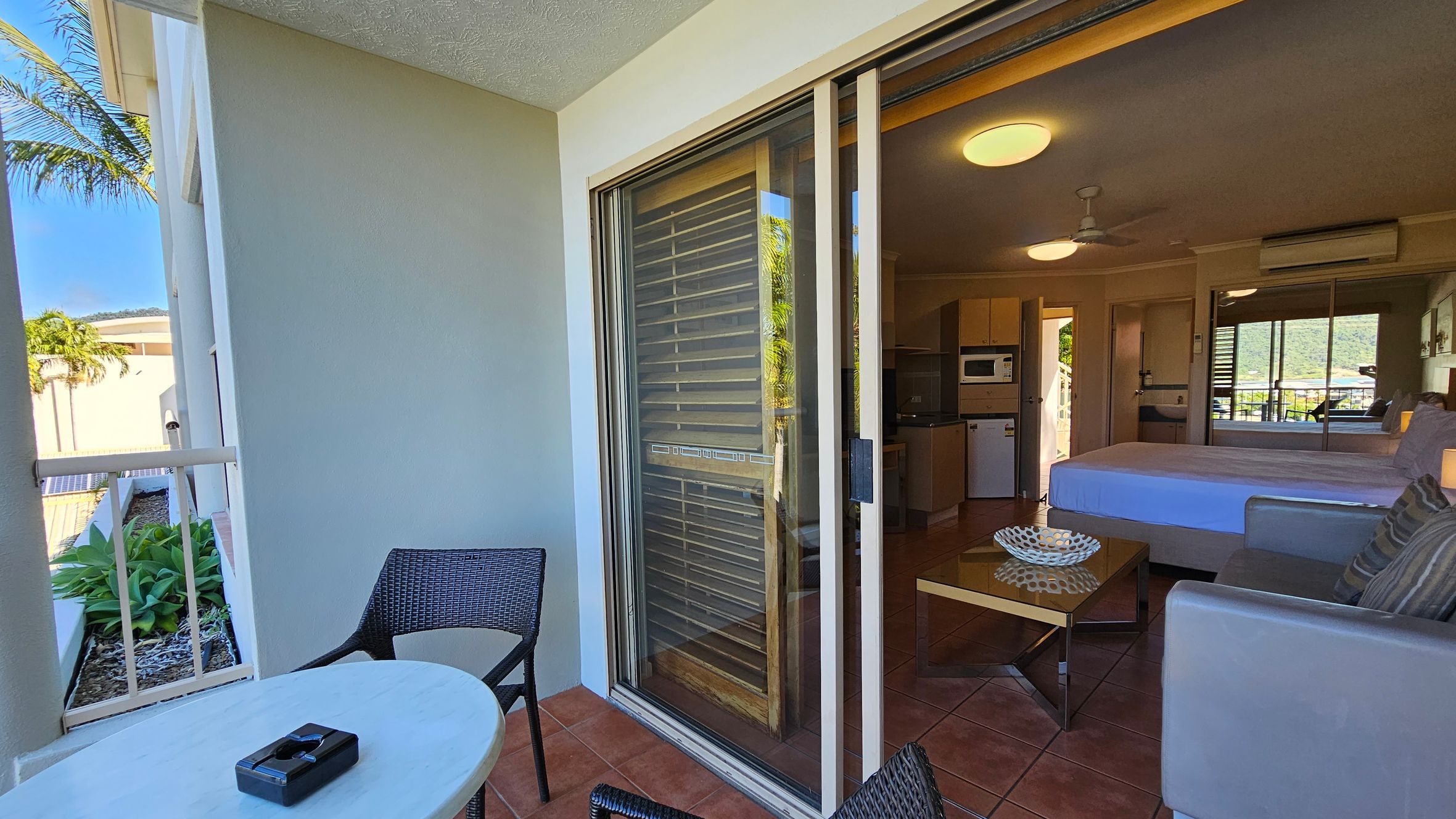 A balcony with terracotta tiles features a small round table and two wicker chairs. A sliding glass door leads to an interior room with a bed, a sofa, and a coffee table. The room has a ceiling fan and an air conditioning unit. A kitchenette with a microwave and a small fridge is visible. Outside, there are green plants and a partial view of a palm tree. A balcony with terracotta tiles features a small round table and two wicker chairs. A sliding glass door leads to an interior room with a bed, a sofa, and a coffee table. The room has a ceiling fan and an air conditioning unit. A kitchenette with a microwave and a small fridge is visible. Outside, there are green plants and a partial view of a palm tree.