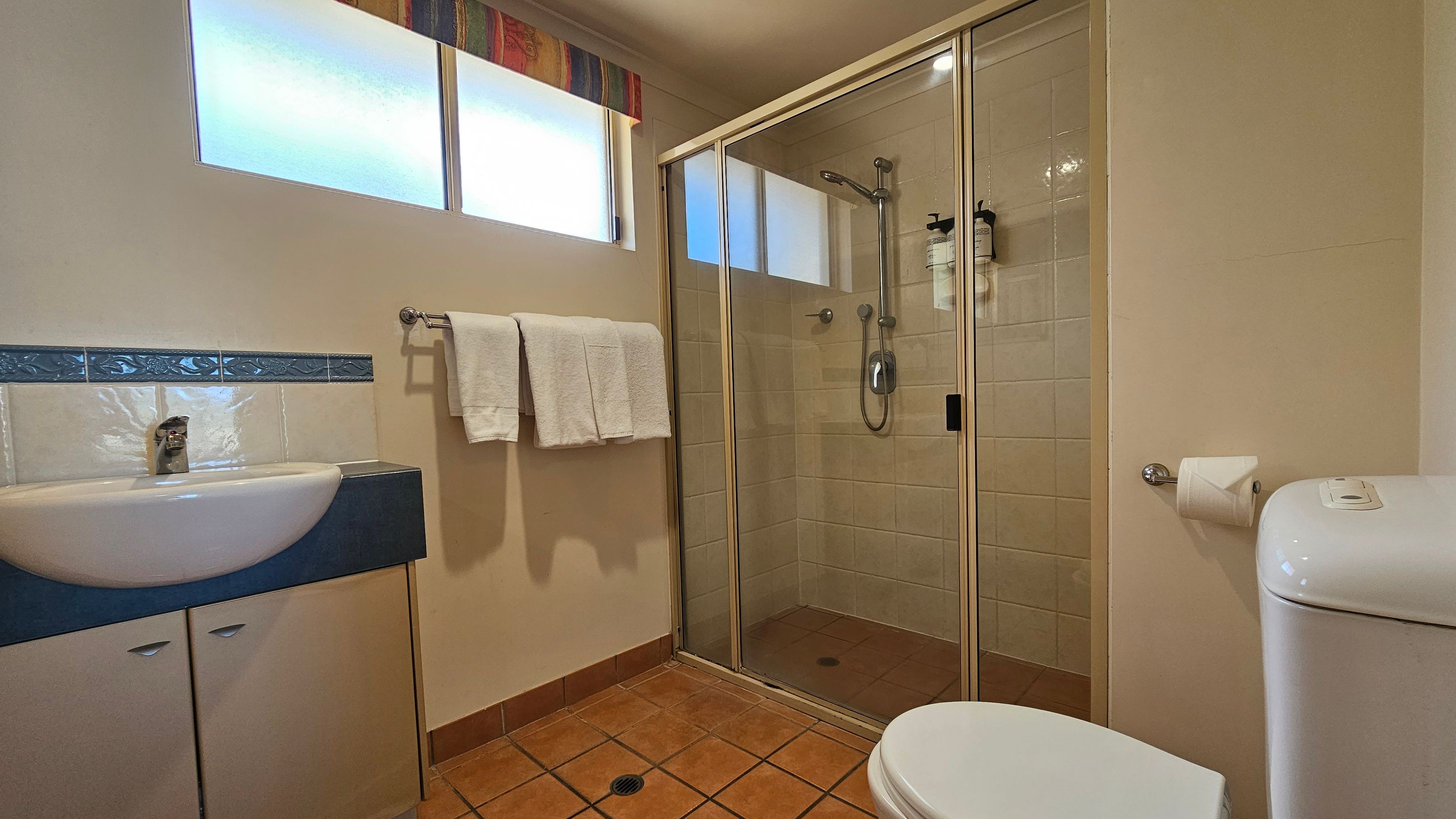 A bathroom with a tiled floor and cream walls. On the left, there's a sink with a blue countertop and a cabinet below, with a decorative blue tile border above. A towel rack with white towels is mounted on the wall. In the centre, a glass-enclosed shower with a sliding door and mounted dispensers. On the right, a toilet with a roll of toilet paper on a holder. Above, frosted windows with a colourful valance allow light in. A bathroom with a tiled floor and cream walls. On the left, there's a sink with a blue countertop and a cabinet below, with a decorative blue tile border above. A towel rack with white towels is mounted on the wall. In the centre, a glass-enclosed shower with a sliding door and mounted dispensers. On the right, a toilet with a roll of toilet paper on a holder. Above, frosted windows with a colourful valance allow light in.