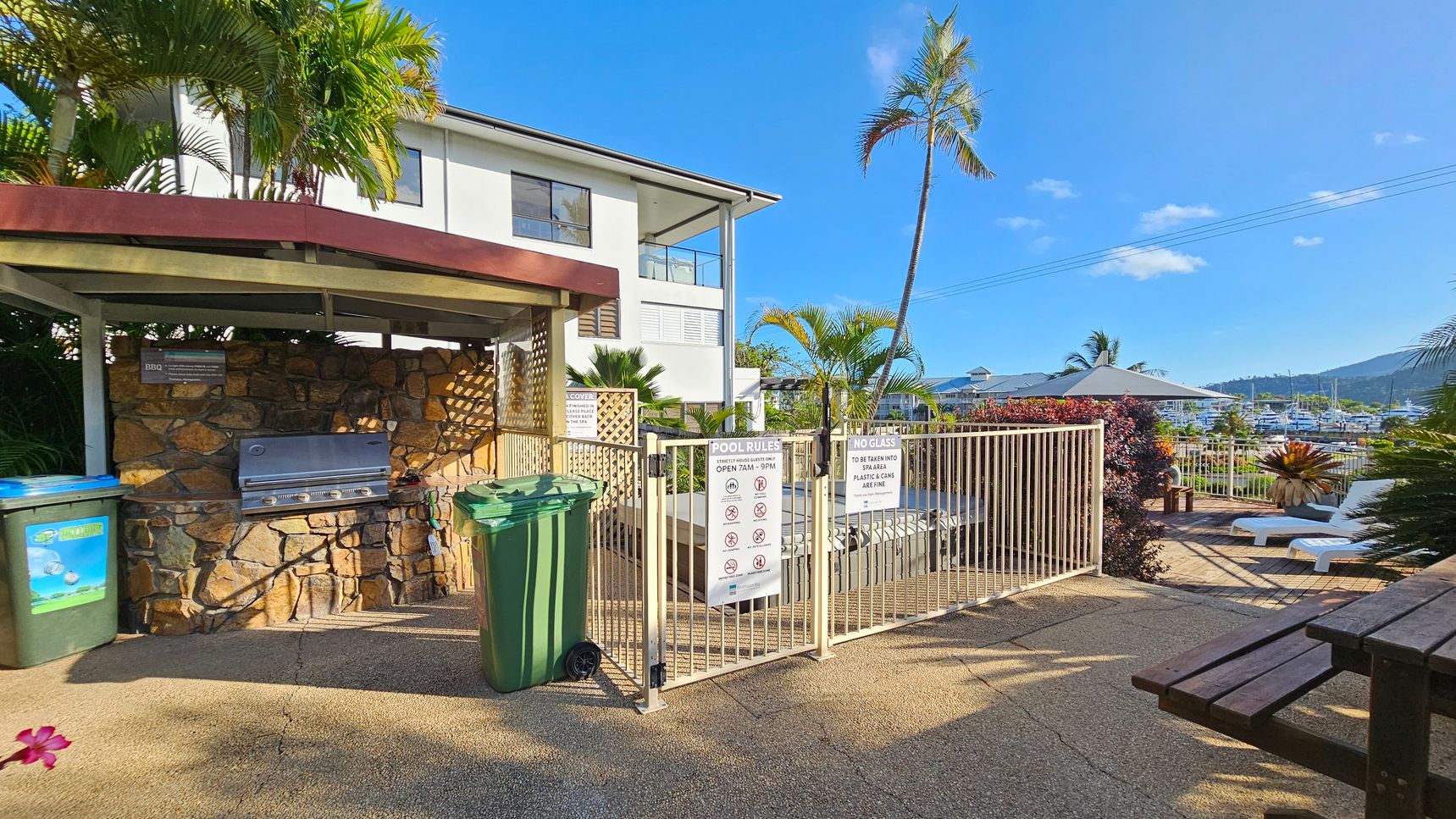 A pool area with a stone barbecue on the left, sheltered by a small roof. Two green bins are nearby. A white building with large windows is in the background. A gated entrance with pool rules is in the centre, leading to a deck with white sun loungers and a large umbrella. Palm trees and lush plants surround the area. In the distance, a marina with boats is visible, set against a backdrop of hills. A wooden bench is in the foreground on the right. A pool area with a stone barbecue on the left, sheltered by a small roof. Two green bins are nearby. A white building with large windows is in the background. A gated entrance with pool rules is in the centre, leading to a deck with white sun loungers and a large umbrella. Palm trees and lush plants surround the area. In the distance, a marina with boats is visible, set against a backdrop of hills. A wooden bench is in the foreground on the right.