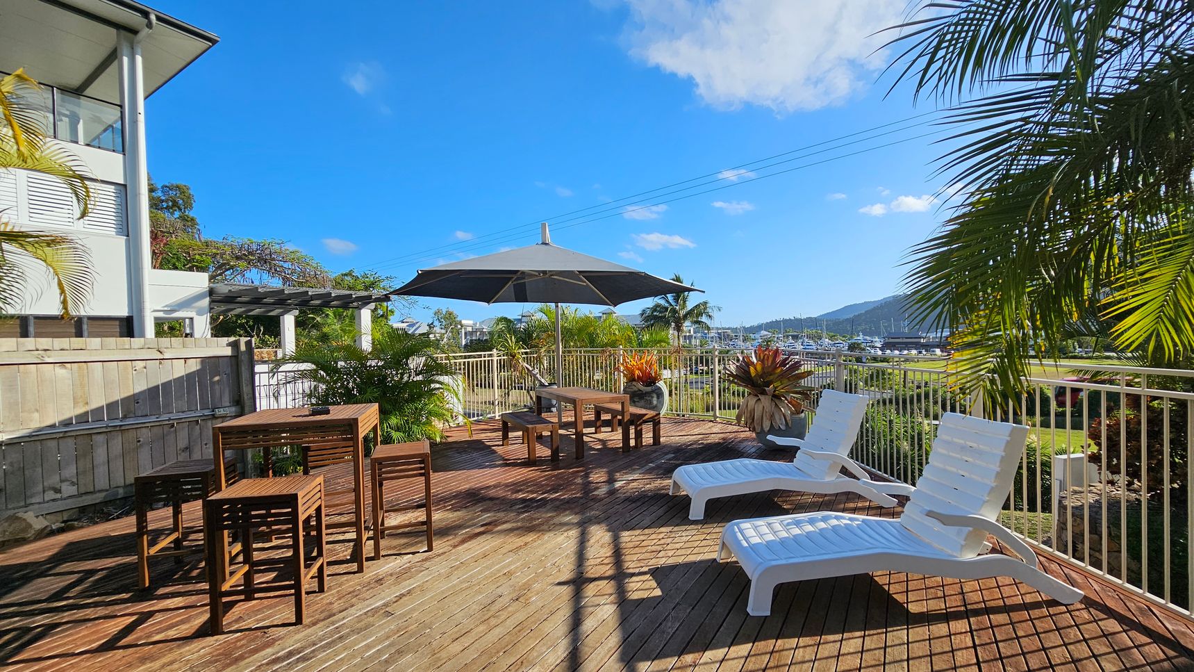 A wooden deck with two white sun loungers on the right and a wooden table with stools on the left. In the centre, a large black umbrella provides shade over a seating area. Potted plants are scattered around, and a metal railing encloses the space. In the background, there are views of a marina with boats and distant hills. A modern building with a wooden fence is on the left, and palm fronds partially frame the scene. A wooden deck with two white sun loungers on the right and a wooden table with stools on the left. In the centre, a large black umbrella provides shade over a seating area. Potted plants are scattered around, and a metal railing encloses the space. In the background, there are views of a marina with boats and distant hills. A modern building with a wooden fence is on the left, and palm fronds partially frame the scene.