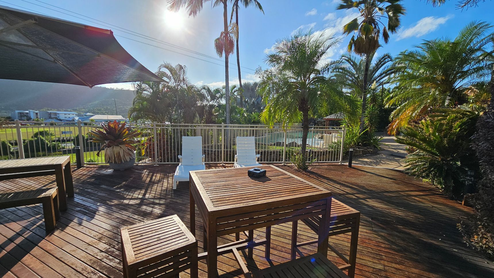 Wooden deck with a table and benches in the foreground. Two white sun loungers are positioned near a metal railing. A large potted plant is on the left. Palm trees and lush greenery are in the background, with a glimpse of a pool area. A sunshade partially covers the deck on the left. The sun is shining brightly, casting shadows on the deck. Wooden deck with a table and benches in the foreground. Two white sun loungers are positioned near a metal railing. A large potted plant is on the left. Palm trees and lush greenery are in the background, with a glimpse of a pool area. A sunshade partially covers the deck on the left. The sun is shining brightly, casting shadows on the deck.