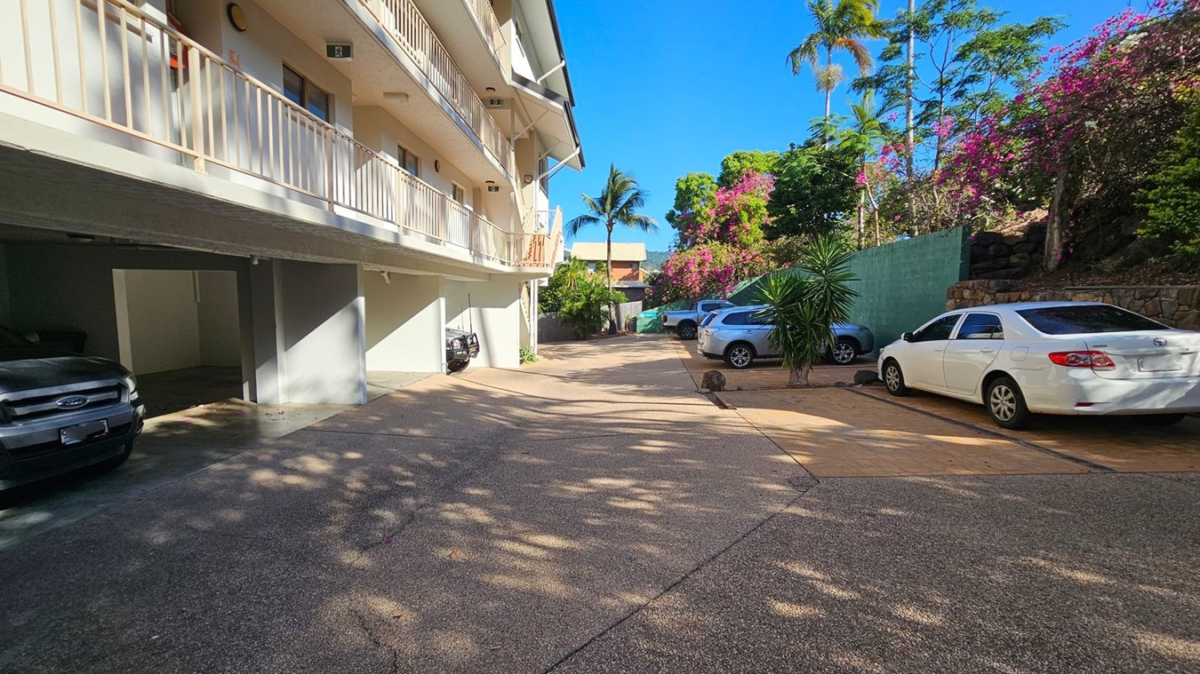 A parking area with several cars is visible. On the left, a building with a light-coloured exterior and a balcony runs along the side. Below the balcony, there are covered parking spaces with cars parked. On the right, a white car is parked near a green fence, with flowering bushes and trees behind it. The ground is paved, and shadows from the trees create patterns on the surface. A palm tree is visible in the background. A parking area with several cars is visible. On the left, a building with a light-coloured exterior and a balcony runs along the side. Below the balcony, there are covered parking spaces with cars parked. On the right, a white car is parked near a green fence, with flowering bushes and trees behind it. The ground is paved, and shadows from the trees create patterns on the surface. A palm tree is visible in the background.