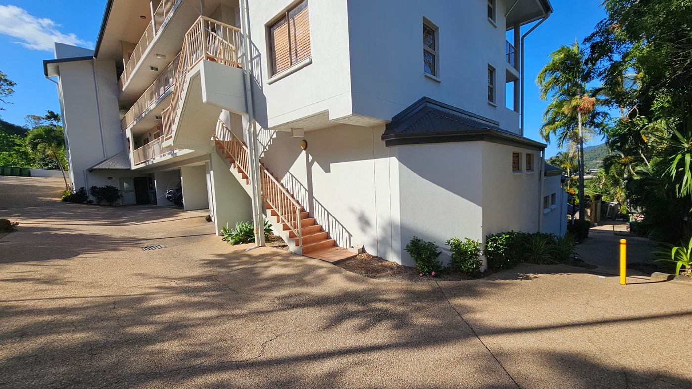 A multi-storey building with white walls and a grey roof is on the right. A staircase with a metal railing leads up to the first floor. The building has several windows with shutters. On the left, there is a driveway with a parked car under a covered area. The ground is paved, and there are small plants and shrubs along the building's base. In the background, there are palm trees and a view of distant hills. Shadows of trees are cast on the ground. A multi-storey building with white walls and a grey roof is on the right. A staircase with a metal railing leads up to the first floor. The building has several windows with shutters. On the left, there is a driveway with a parked car under a covered area. The ground is paved, and there are small plants and shrubs along the building's base. In the background, there are palm trees and a view of distant hills. Shadows of trees are cast on the ground.