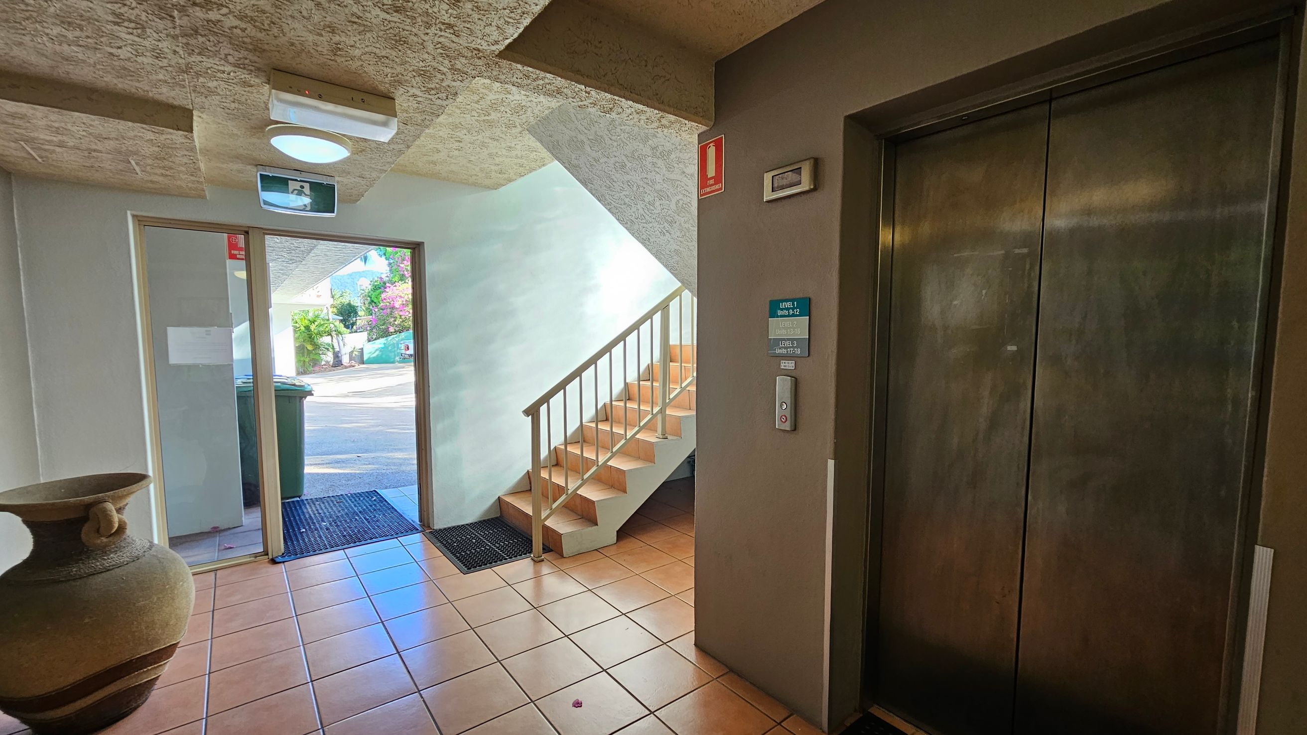 A hallway with a tiled floor and textured ceiling. On the right, there is a closed lift with a control panel and signage indicating floor levels. In the centre, a staircase with a metal railing leads upwards. On the left, a large decorative pot sits near a glass door, which opens to the outside. A green wheelie bin is visible through the door. A light fixture and an exit sign are mounted on the ceiling. A hallway with a tiled floor and textured ceiling. On the right, there is a closed lift with a control panel and signage indicating floor levels. In the centre, a staircase with a metal railing leads upwards. On the left, a large decorative pot sits near a glass door, which opens to the outside. A green wheelie bin is visible through the door. A light fixture and an exit sign are mounted on the ceiling.