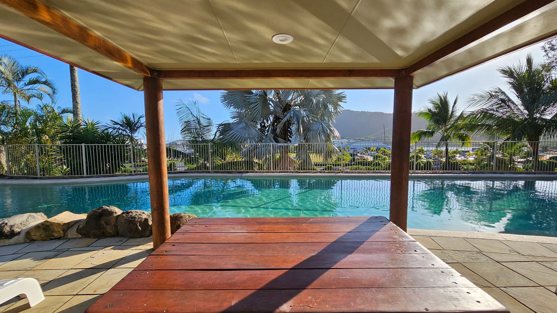 A gated pool area with a corrugated metal roofed shelter in the centre. The gate has signs displaying pool rules and a notice about no glass allowed. Lush greenery, including palm trees and various plants, surrounds the area. Beyond the gate, a glimpse of the pool is visible. The ground is paved with small stones, and the sun is shining brightly, casting shadows. A gated pool area with a corrugated metal roofed shelter in the centre. The gate has signs displaying pool rules and a notice about no glass allowed. Lush greenery, including palm trees and various plants, surrounds the area. Beyond the gate, a glimpse of the pool is visible. The ground is paved with small stones, and the sun is shining brightly, casting shadows.