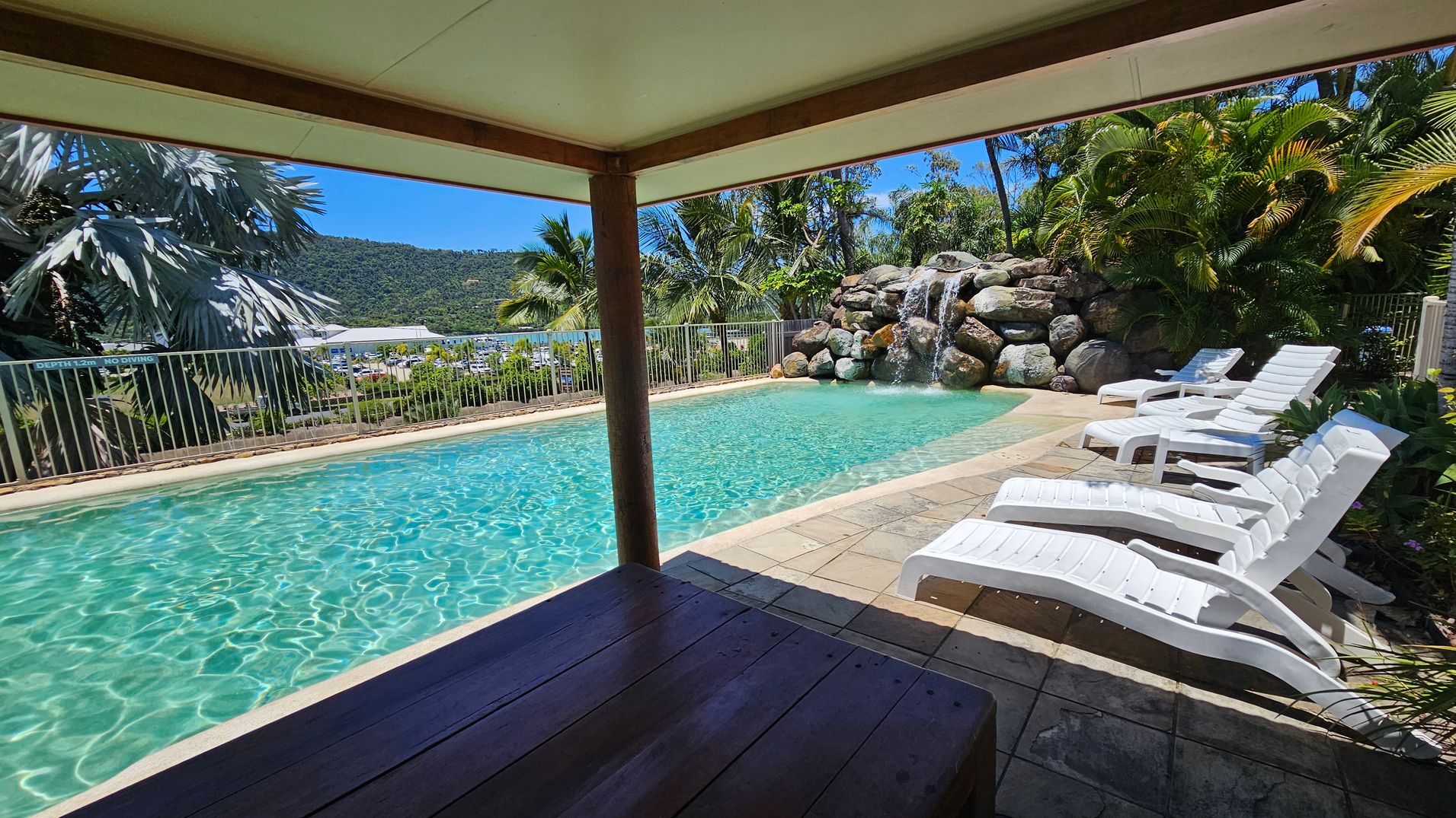A swimming pool with clear water is surrounded by a tiled deck. On the right, several white sun loungers are lined up. In the background, a rock formation with a waterfall is visible, along with lush green palm trees and foliage. A metal fence runs along the left side, with a view of a marina and hills in the distance. The foreground shows a shaded area with a wooden table. A swimming pool with clear water is surrounded by a tiled deck. On the right, several white sun loungers are lined up. In the background, a rock formation with a waterfall is visible, along with lush green palm trees and foliage. A metal fence runs along the left side, with a view of a marina and hills in the distance. The foreground shows a shaded area with a wooden table.