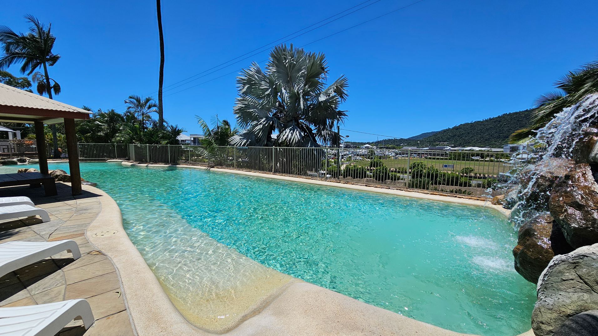 A swimming pool with clear blue water is in the centre, featuring a small waterfall on the right cascading over rocks. On the left, there are several white sun loungers on a tiled patio under a shaded area. In the background, there are palm trees and a metal fence, with a view of a distant hillside and some buildings. The sky is clear and blue. A swimming pool with clear blue water is in the centre, featuring a small waterfall on the right cascading over rocks. On the left, there are several white sun loungers on a tiled patio under a shaded area. In the background, there are palm trees and a metal fence, with a view of a distant hillside and some buildings. The sky is clear and blue.