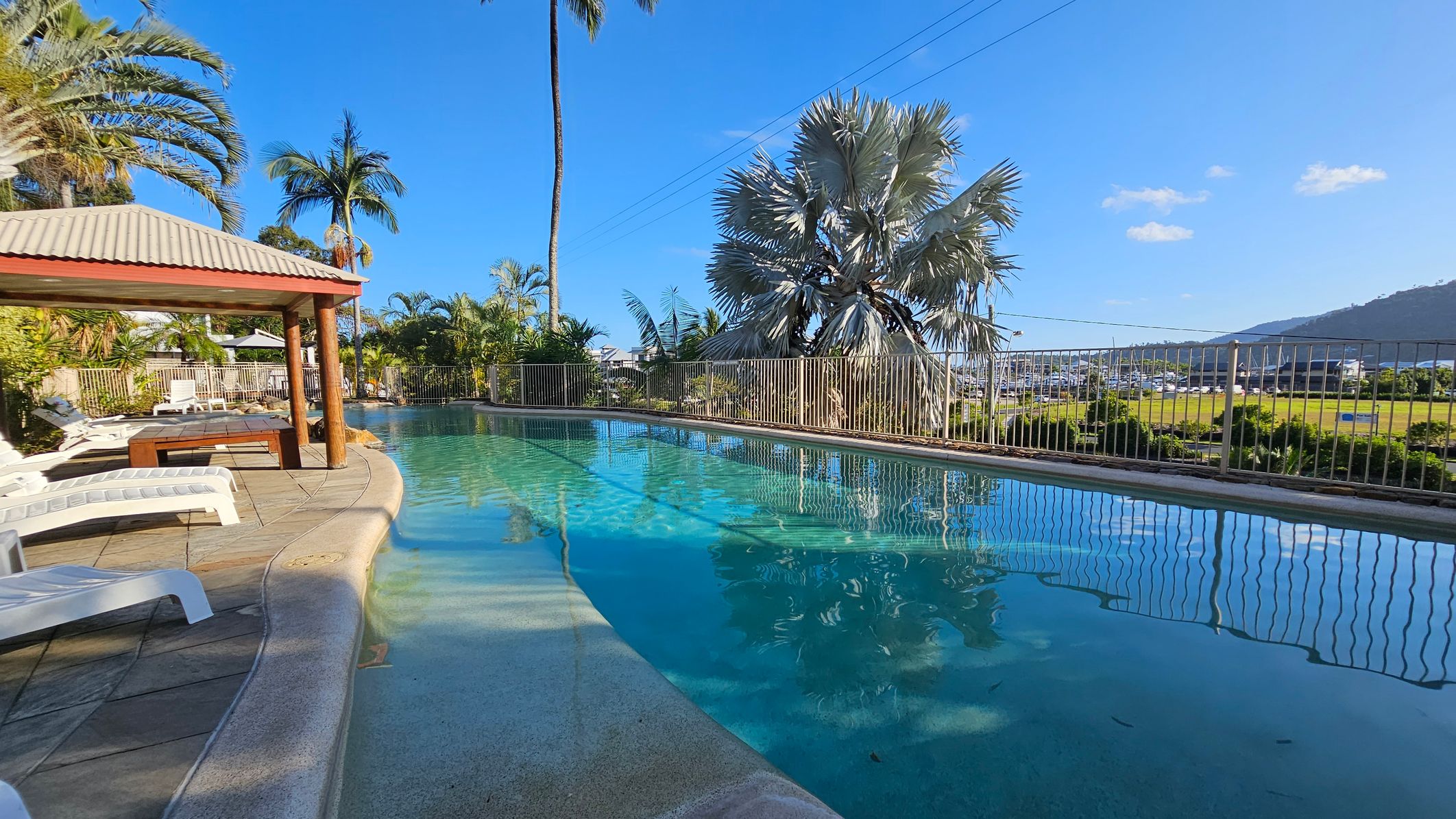 A swimming pool with clear blue water is on the right, bordered by a metal fence. On the left, there is a shaded area with a wooden structure and several white sun loungers on a tiled floor. Tall palm trees and lush greenery surround the pool area. In the background, there is a view of a grassy field and a marina with boats, set against a backdrop of hills. A swimming pool with clear blue water is on the right, bordered by a metal fence. On the left, there is a shaded area with a wooden structure and several white sun loungers on a tiled floor. Tall palm trees and lush greenery surround the pool area. In the background, there is a view of a grassy field and a marina with boats, set against a backdrop of hills.