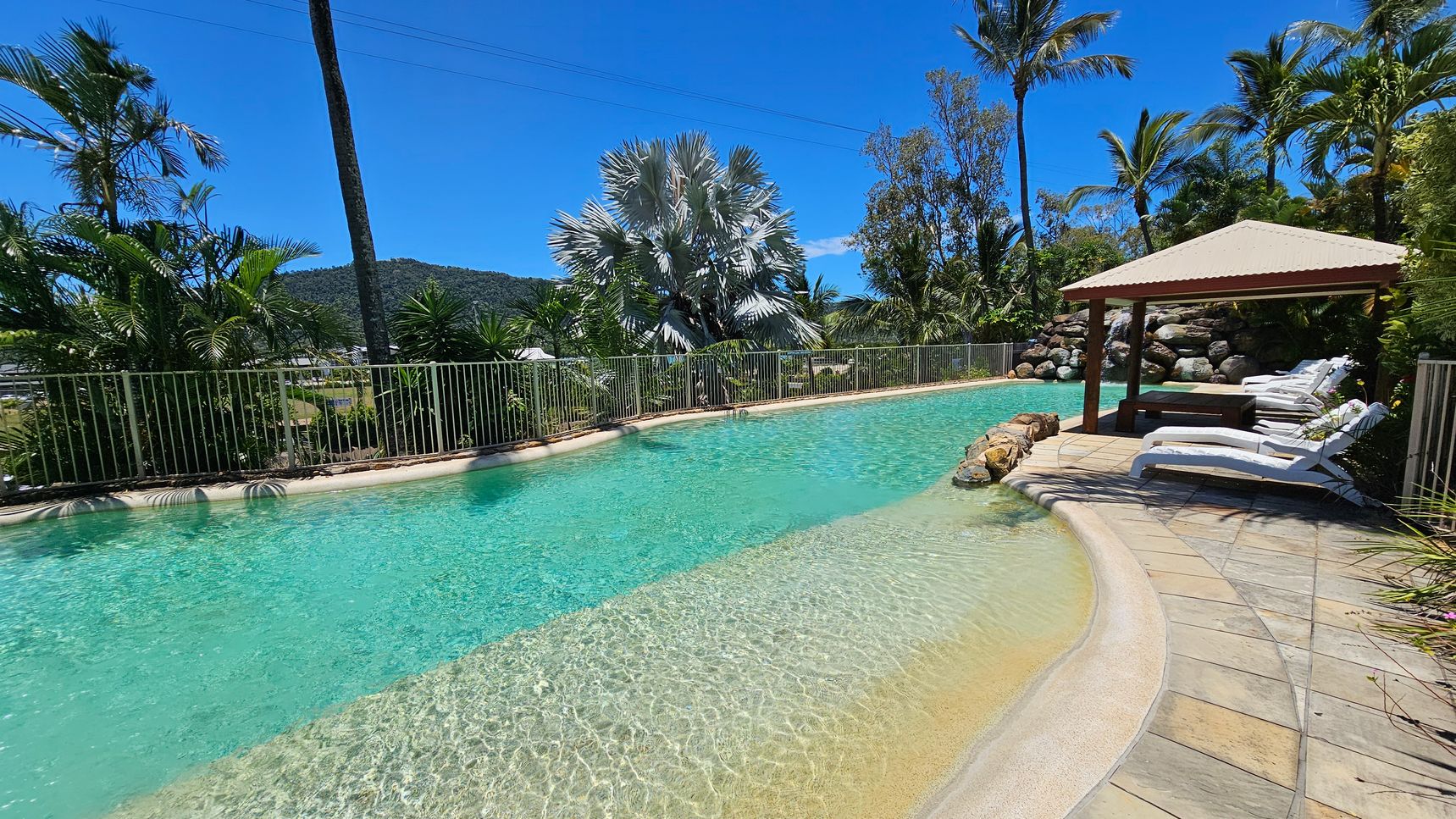 A swimming pool with clear turquoise water is surrounded by a stone-tiled deck. On the right, several white sun loungers are positioned under a wooden gazebo with a corrugated roof. In the background, lush tropical plants and palm trees line the perimeter, with a metal fence separating the pool area from the greenery. A hill covered in vegetation is visible in the distance. A swimming pool with clear turquoise water is surrounded by a stone-tiled deck. On the right, several white sun loungers are positioned under a wooden gazebo with a corrugated roof. In the background, lush tropical plants and palm trees line the perimeter, with a metal fence separating the pool area from the greenery. A hill covered in vegetation is visible in the distance.