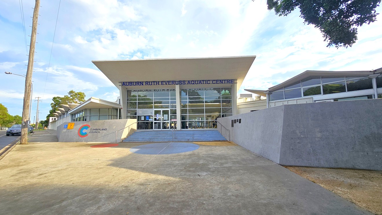 Concreted open space in front of Auburn Ruth Everuss Aquatic Centre. The facility is a large white brick building with glass elements on the front wall. The concrete wall on the left with green, blue, and red color lines forming a C beside ‘CUMBERLAND COUNCIL’ and the right concrete wall with black bar tiles distinguishes the arrival area. In the middle are the concrete stairs with metal handrails that lead to the glass entrance wall with a blue ‘AUBURN RUTH EVERUSS AQUATIC CENTRE’ detailed. Concreted open space in front of Auburn Ruth Everuss Aquatic Centre. The facility is a large white brick building with glass elements on the front wall. The concrete wall on the left with green, blue, and red color lines forming a C beside ‘CUMBERLAND COUNCIL’ and the right concrete wall with black bar tiles distinguishes the arrival area. In the middle are the concrete stairs with metal handrails that lead to the glass entrance wall with a blue ‘AUBURN RUTH EVERUSS AQUATIC CENTRE’ detailed.