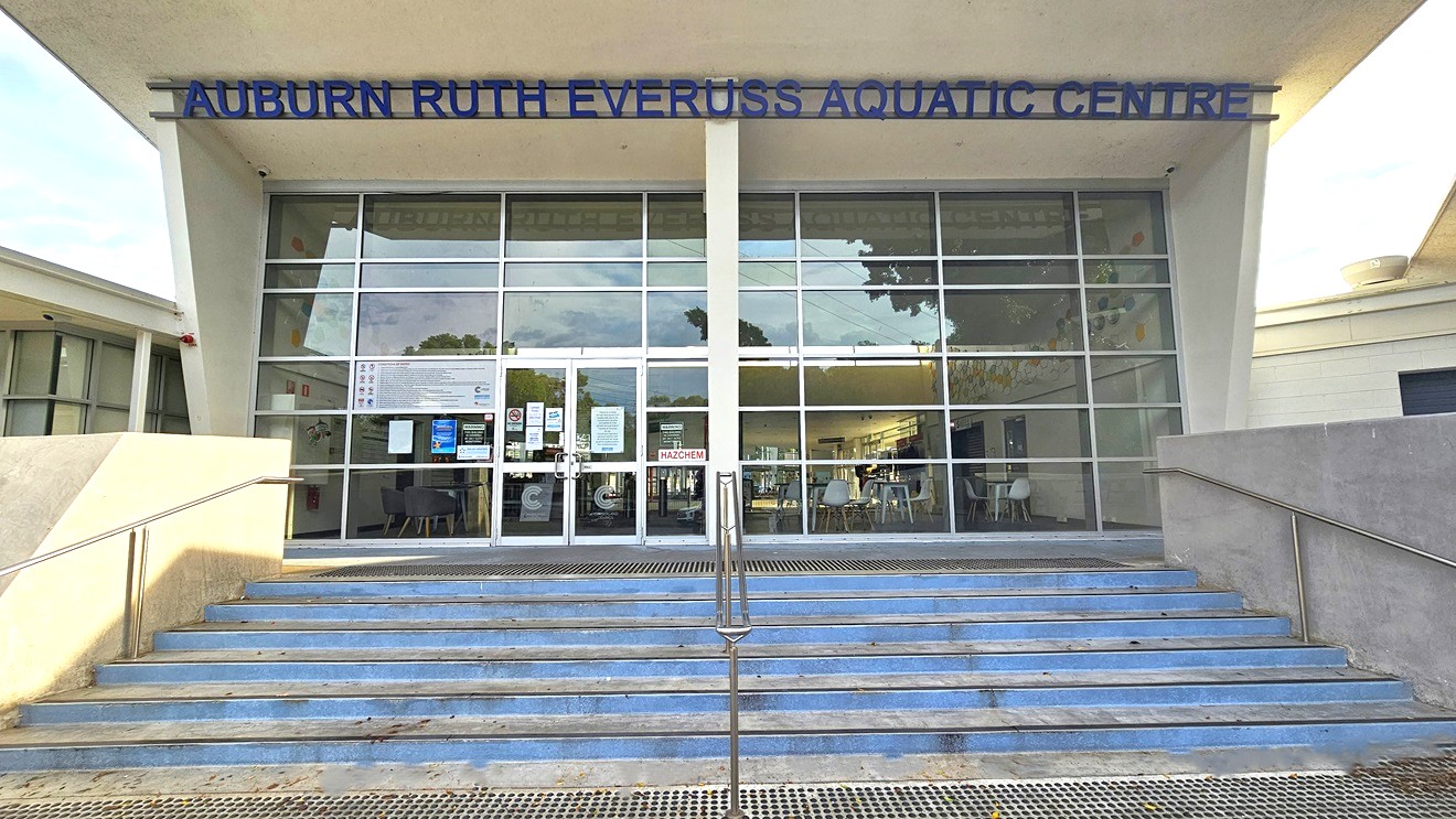 The stairs with metal handrails have light blue painted steps. At the bottom and top of the stairs are metal gratings for water drainage. The entrance door is on the left side, surrounded by varying kinds, colors, and sizes of informational signs and adverts. Two light gray lines forming a C are on the bottom of the glass entrance doors. On the top of the front wall is a blue ‘AUBURN RUTH EVERUSS AQUATIC CENTRE.’ Seen through the glass door and wall are the chairs, tables, and doors inside. The stairs with metal handrails have light blue painted steps. At the bottom and top of the stairs are metal gratings for water drainage. The entrance door is on the left side, surrounded by varying kinds, colors, and sizes of informational signs and adverts. Two light gray lines forming a C are on the bottom of the glass entrance doors. On the top of the front wall is a blue ‘AUBURN RUTH EVERUSS AQUATIC CENTRE.’ Seen through the glass door and wall are the chairs, tables, and doors inside.