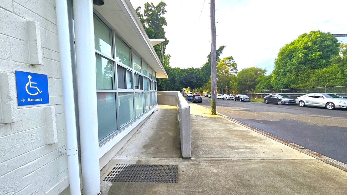 Ramp access marked by a blue square sign with a white image of a person in a wheelchair placed on the white brick wall. On the sides of the sign are three bricks protruding from the wall. Near it are small and large white pipes connected from the roof to the floor. Next to the pipes are blurry square glass windows with white metal frames. The ramp access has stainless steel handrails mounted on the right concrete wall. Several cars of different colors are parked on each side of the road. Ramp access marked by a blue square sign with a white image of a person in a wheelchair placed on the white brick wall. On the sides of the sign are three bricks protruding from the wall. Near it are small and large white pipes connected from the roof to the floor. Next to the pipes are blurry square glass windows with white metal frames. The ramp access has stainless steel handrails mounted on the right concrete wall. Several cars of different colors are parked on each side of the road.