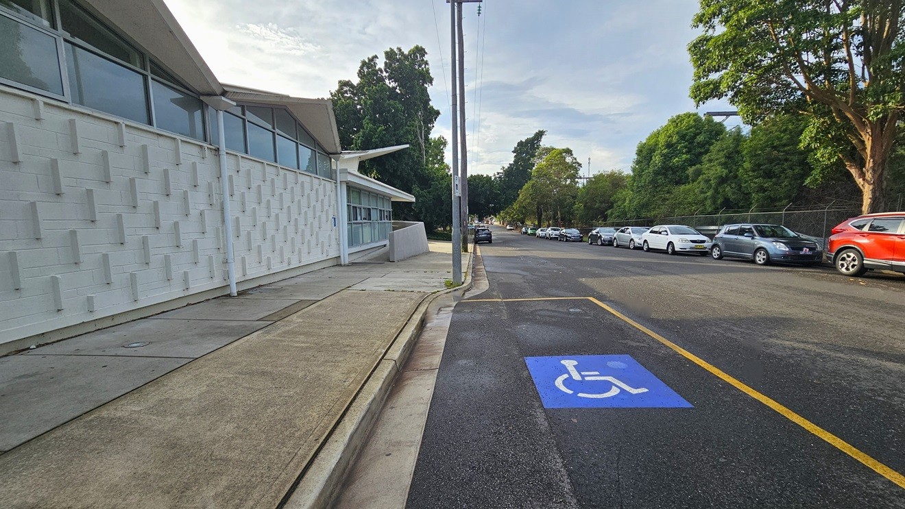 Further to the back of the left concrete wall with ramp access is the accessible car parking space for persons with mobility disabilities. The accessible car parking space has a blue image with a white wheelchair placed on the road and is separated by a yellow rectangular line. The white wall of the building has several protruding rectangular, narrow white bricks, while the upper part is made of glass windows with white metal frames. Further to the back of the left concrete wall with ramp access is the accessible car parking space for persons with mobility disabilities. The accessible car parking space has a blue image with a white wheelchair placed on the road and is separated by a yellow rectangular line. The white wall of the building has several protruding rectangular, narrow white bricks, while the upper part is made of glass windows with white metal frames.