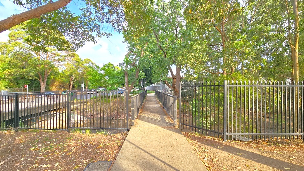 Concrete pedestrian bridge from the right of Auburn Ruth Everrus Aquatic Centre with a black metal fence on each side. Under the bridge is a waterway that starts from the left and continues to the right and is marked by a higher black metal fence. Tall trees with abundant foliage offer significant shade to this walkway. Concrete pedestrian bridge from the right of Auburn Ruth Everrus Aquatic Centre with a black metal fence on each side. Under the bridge is a waterway that starts from the left and continues to the right and is marked by a higher black metal fence. Tall trees with abundant foliage offer significant shade to this walkway.