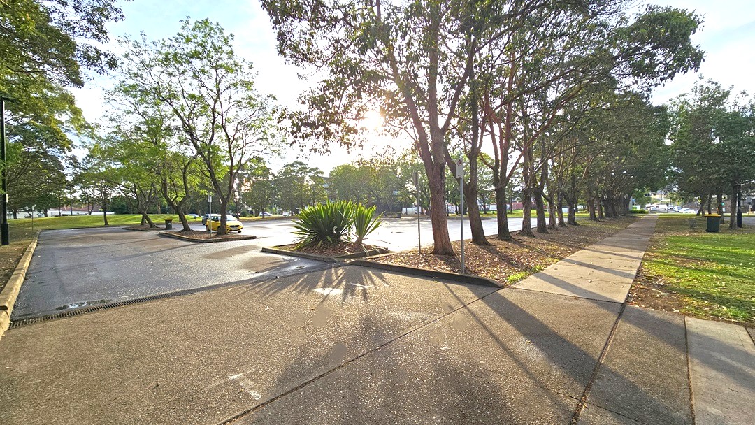 Extensive car park from the right side of the facility. Around the car park are trees lined on the edge of the garden. A No Parking sign with a red arrow at the bottom pointing towards the entry space to the extensive car park can be found on the right of the main entrance. Near it are two green plants from the palm family. Large grey trash bins with yellow covers are placed inside the car park and on the side of the concrete walkway. Extensive car park from the right side of the facility. Around the car park are trees lined on the edge of the garden. A No Parking sign with a red arrow at the bottom pointing towards the entry space to the extensive car park can be found on the right of the main entrance. Near it are two green plants from the palm family. Large grey trash bins with yellow covers are placed inside the car park and on the side of the concrete walkway.