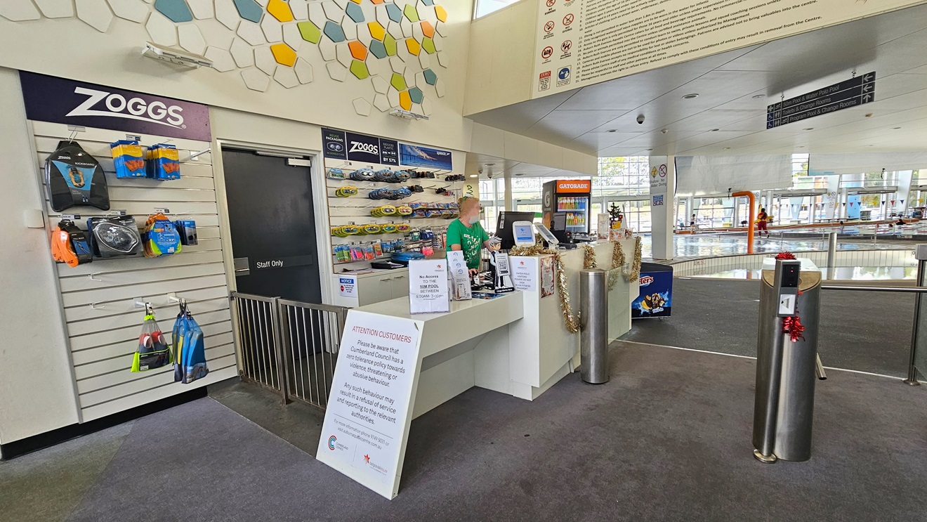 Closer view of the reception area. A small retail area that displays goggles and swimming accessories is visible behind the receptionist. Posters are placed above the low-height counter and beside the gated entrance of the reception desk. Further ahead, there is a Gatorade display placed near an ice cream freezer. Closer view of the reception area. A small retail area that displays goggles and swimming accessories is visible behind the receptionist. Posters are placed above the low-height counter and beside the gated entrance of the reception desk. Further ahead, there is a Gatorade display placed near an ice cream freezer.