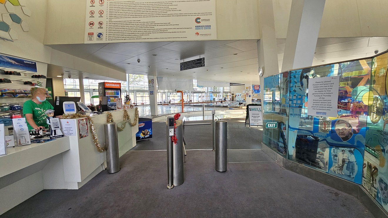Closer view of the gated barrier leading inside the facility. The glass walls on the right side feature a collage of images and promotional materials. A signage and an exit sign are also affixed on the glass walls. A red Christmas decor is placed in the center of the gated barrier. On the left, golden Christmas decor hangs on the front of the reception desk. Further ahead, a directional sign is situated above the ceiling. Closer view of the gated barrier leading inside the facility. The glass walls on the right side feature a collage of images and promotional materials. A signage and an exit sign are also affixed on the glass walls. A red Christmas decor is placed in the center of the gated barrier. On the left, golden Christmas decor hangs on the front of the reception desk. Further ahead, a directional sign is situated above the ceiling.