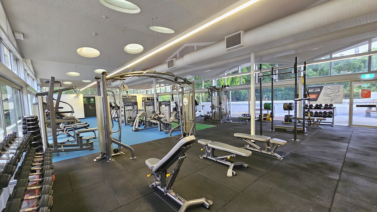 On the left, dumbbells are arranged near the wall, and lat pulldown machines are set up alongside. Bench chairs are positioned in the middle. Smith Machines and stability balls are placed to the right. Further in, more machines are arranged on the blue flooring. The weights area floor is covered with black rubber tiles. The ceiling is white, featuring LED and fluorescent lights with large aircon vents. The right wall consists of floor-to-ceiling glass with posters, offering a view outside. On the left, dumbbells are arranged near the wall, and lat pulldown machines are set up alongside. Bench chairs are positioned in the middle. Smith Machines and stability balls are placed to the right. Further in, more machines are arranged on the blue flooring. The weights area floor is covered with black rubber tiles. The ceiling is white, featuring LED and fluorescent lights with large aircon vents. The right wall consists of floor-to-ceiling glass with posters, offering a view outside.