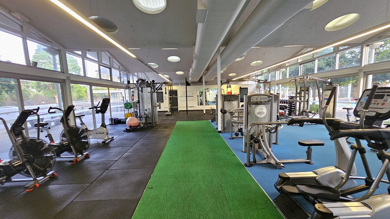 Spin machines and Smith Machines are arranged on the left, alongside lateral machines and others in the middle. Further in, free weights and Lat Pulldown machines are placed. The surrounding wall is made of glass, offering a view outside. The floor is a combination of black rubber tiles, green, and blue tiles. The ceiling is all white with LED and fluorescent lights, along with large air conditioning vents. Spin machines and Smith Machines are arranged on the left, alongside lateral machines and others in the middle. Further in, free weights and Lat Pulldown machines are placed. The surrounding wall is made of glass, offering a view outside. The floor is a combination of black rubber tiles, green, and blue tiles. The ceiling is all white with LED and fluorescent lights, along with large air conditioning vents.