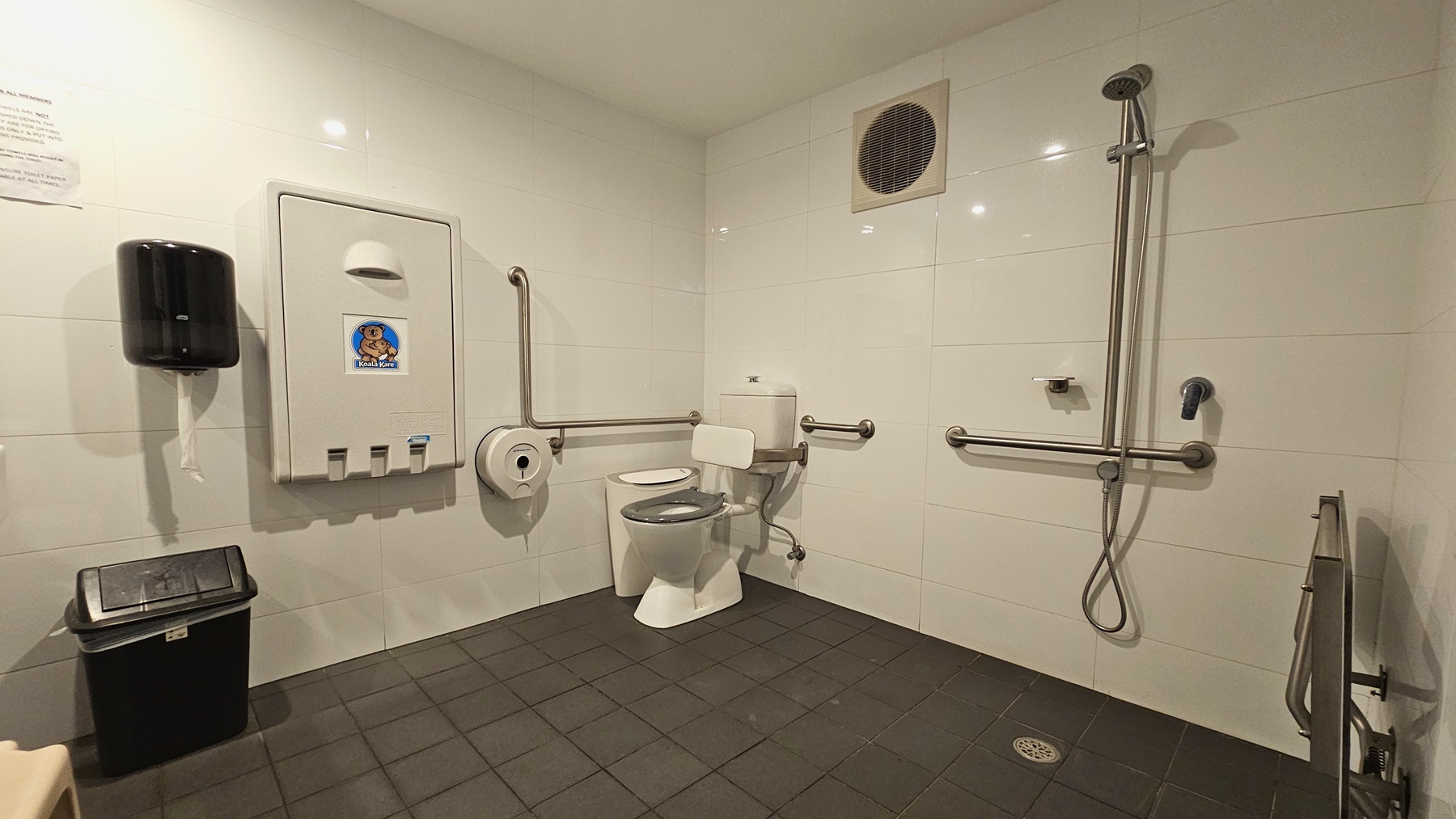 Within the accessible bathroom, a toilet bowl equipped with hand grabs is positioned on the left, accompanied by a rubbish bin. On the left wall, there's a tissue dispenser, a foldable change table, and another rubbish bin. To the right, a shower with handrails is present, and a foldable bench is positioned against the wall. The floor is covered with grey tiles, and the walls have an exhaust fan, while the ceiling is white. Within the accessible bathroom, a toilet bowl equipped with hand grabs is positioned on the left, accompanied by a rubbish bin. On the left wall, there's a tissue dispenser, a foldable change table, and another rubbish bin. To the right, a shower with handrails is present, and a foldable bench is positioned against the wall. The floor is covered with grey tiles, and the walls have an exhaust fan, while the ceiling is white.
