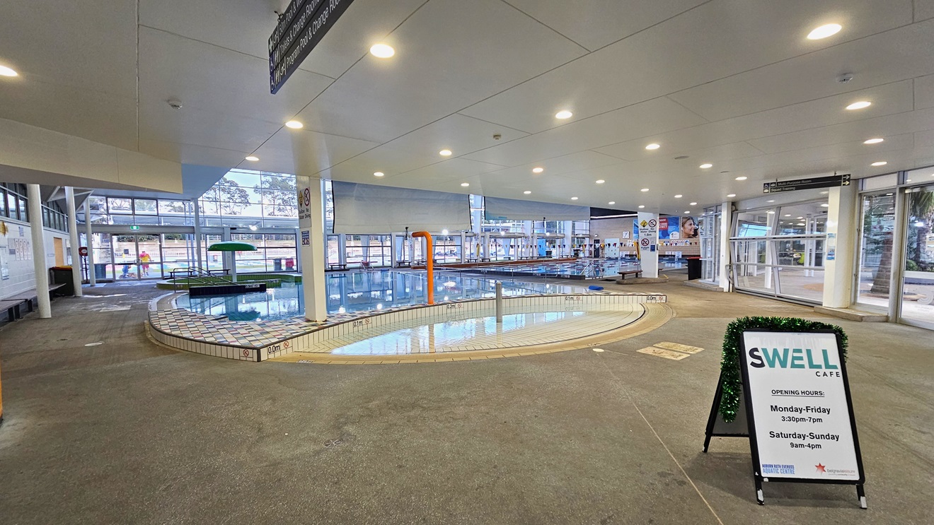 Leisure and Toddler Pool with a beach entry, an umbrella fountain, and stairs with handrails. To the right is a free-standing banner for the Cafe equipped with a Christmas garland. To the far right is another pool. The walls in the middle and the right are made of glass. Banners, safety warning signs, and directional signs are visible. The floor has concrete tiles, and the ceiling is all white with LED lights. Leisure and Toddler Pool with a beach entry, an umbrella fountain, and stairs with handrails. To the right is a free-standing banner for the Cafe equipped with a Christmas garland. To the far right is another pool. The walls in the middle and the right are made of glass. Banners, safety warning signs, and directional signs are visible. The floor has concrete tiles, and the ceiling is all white with LED lights.