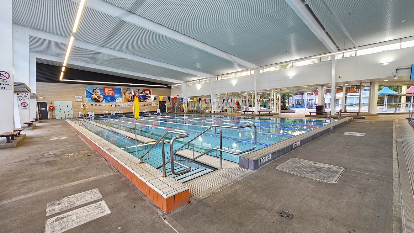 The 25m pool is equipped with steps and a ramp with handrails for entry. Truncated domes are installed before the steps. The pool is surrounded by a raised pool deck with markings denoting the pool depth. Floating ropes and colorful buntings run across the pool. Bench chairs are arranged on all sides of the pool. The middle wall features cream bricks, with large banners and security warnings affixed. The ceiling features a corrugated design with exposed metal beams and fluorescent lights. The 25m pool is equipped with steps and a ramp with handrails for entry. Truncated domes are installed before the steps. The pool is surrounded by a raised pool deck with markings denoting the pool depth. Floating ropes and colorful buntings run across the pool. Bench chairs are arranged on all sides of the pool. The middle wall features cream bricks, with large banners and security warnings affixed. The ceiling features a corrugated design with exposed metal beams and fluorescent lights.