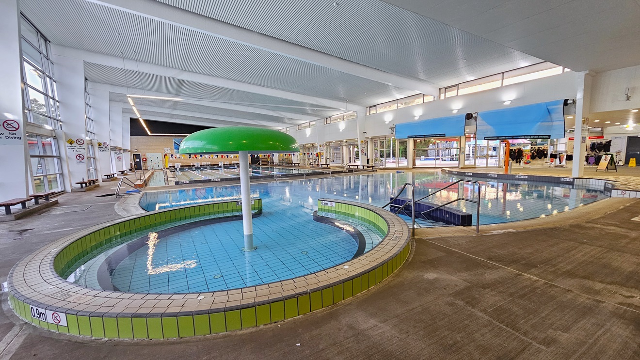 The Leisure and Toddler Pool has a stepped entry with handrails and a green umbrella fountain. To the right, banners and swimwear for sale are displayed near the pool deck. A rolled blue mat is positioned overhead. Adjacent is the 25m pool, equipped with a ramp and steps with handrails. Bench chairs are arranged on the left wall against the glass window. The ceiling features a white corrugated design with exposed metal beams and fluorescent lights. The Leisure and Toddler Pool has a stepped entry with handrails and a green umbrella fountain. To the right, banners and swimwear for sale are displayed near the pool deck. A rolled blue mat is positioned overhead. Adjacent is the 25m pool, equipped with a ramp and steps with handrails. Bench chairs are arranged on the left wall against the glass window. The ceiling features a white corrugated design with exposed metal beams and fluorescent lights.