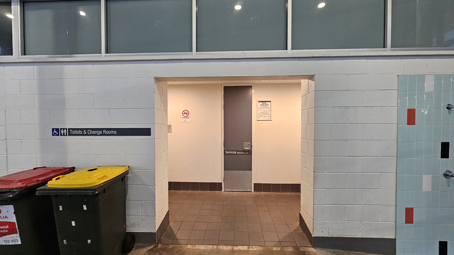 The accessible toilets and change rooms feature a white wall with a brick design. A signage is affixed to the left side of the wall. Two large rubbish bins are on the left. On the right is an outdoor shower area with multi-color brick tile accents. The upper wall features all glass, and the floor has brown tiles. Inside, the grey door is placed between two signages. The accessible toilets and change rooms feature a white wall with a brick design. A signage is affixed to the left side of the wall. Two large rubbish bins are on the left. On the right is an outdoor shower area with multi-color brick tile accents. The upper wall features all glass, and the floor has brown tiles. Inside, the grey door is placed between two signages.