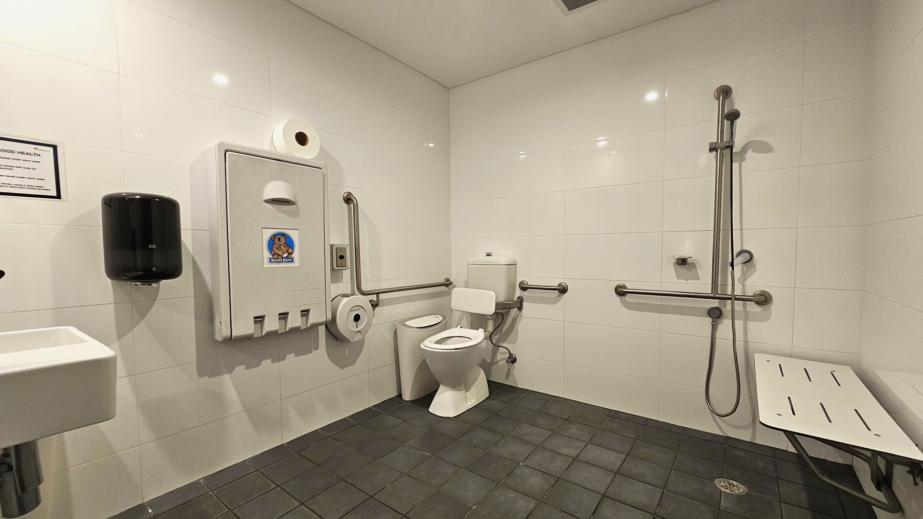 Inside the accessible bathroom, to the left is a toilet bowl equipped with hand grabs. A rubbish bin is placed beside it. A tissue dispenser, a foldable change table, and a liquid hand soap dispenser are mounted on the left wall. A wash basin and a poster are mounted on the wall on the left. To the right is a shower with handrails. A bench is positioned against the right wall. The floor has grey tiles, and the walls are white. Inside the accessible bathroom, to the left is a toilet bowl equipped with hand grabs. A rubbish bin is placed beside it. A tissue dispenser, a foldable change table, and a liquid hand soap dispenser are mounted on the left wall. A wash basin and a poster are mounted on the wall on the left. To the right is a shower with handrails. A bench is positioned against the right wall. The floor has grey tiles, and the walls are white.