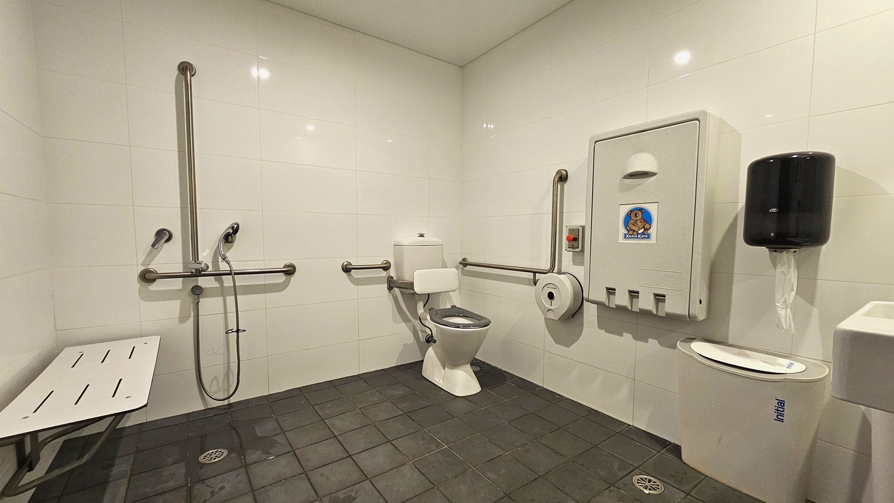 Inside the accessible bathroom, to the right is a toilet bowl equipped with hand grabs. A rubbish bin is placed beside it. A tissue dispenser, a foldable change table, and a liquid hand soap dispenser are mounted on the right wall. A wash basin and a poster are mounted on the wall on the right. To the left is a shower with handrails. A bench is positioned against the left wall. The floor has grey tiles, and the walls and ceiling are white. Inside the accessible bathroom, to the right is a toilet bowl equipped with hand grabs. A rubbish bin is placed beside it. A tissue dispenser, a foldable change table, and a liquid hand soap dispenser are mounted on the right wall. A wash basin and a poster are mounted on the wall on the right. To the left is a shower with handrails. A bench is positioned against the left wall. The floor has grey tiles, and the walls and ceiling are white.