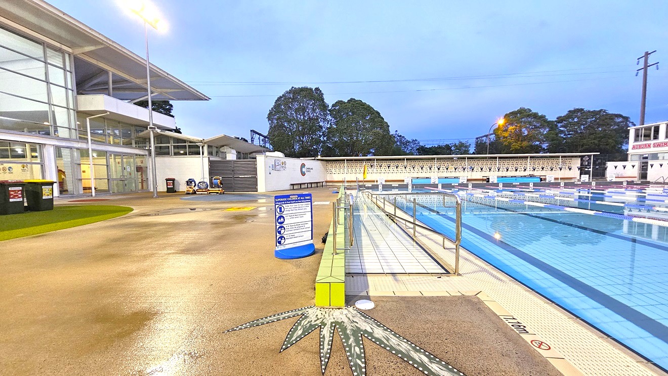 In the middle is the 50m 8-lane lap pool equipped with a ramp and step entry with handrails. Floating ropes and blue buntings run across the pool. Diving boards with covers are aligned on the raised pool deck on the right. Beyond the pool is an extensive tiered seating with shade. To the far left is a two-storey white building. Trees surround the area. In the middle is the 50m 8-lane lap pool equipped with a ramp and step entry with handrails. Floating ropes and blue buntings run across the pool. Diving boards with covers are aligned on the raised pool deck on the right. Beyond the pool is an extensive tiered seating with shade. To the far left is a two-storey white building. Trees surround the area.