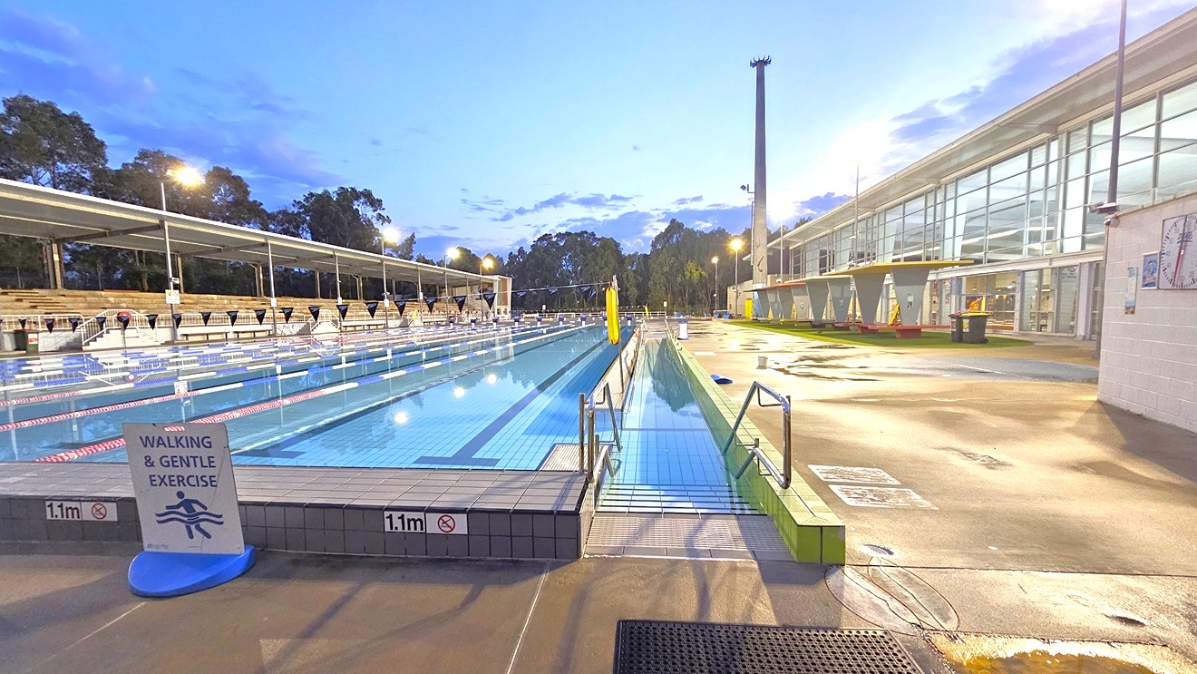 In the middle is the 50m 8-lane lap pool equipped with a ramp and handrails. A free standing banner stands on the pool deck. Floating ropes and blue buntings run across the pool. Truncated domes and a non-slippery mat adorn the ramp entrance. To the left is an extensive tiered seating with shade. To the right is the facility building with glass walls. Shade structures, seating, and rubbish bins are visible in the grass area. In the middle is the 50m 8-lane lap pool equipped with a ramp and handrails. A free standing banner stands on the pool deck. Floating ropes and blue buntings run across the pool. Truncated domes and a non-slippery mat adorn the ramp entrance. To the left is an extensive tiered seating with shade. To the right is the facility building with glass walls. Shade structures, seating, and rubbish bins are visible in the grass area.