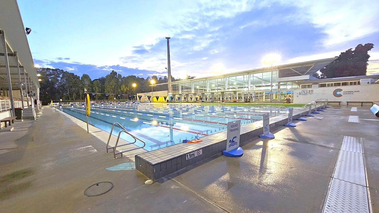 In the middle is the 50m 8-lane lap pool equipped with a ramp and handrails. A free standing banner stands on the pool deck. Floating ropes and blue buntings run across the pool. To the left is an extensive tiered seating with shade. To the right is the facility building with glass walls. Shade structures, seating, and rubbish bins are visible in the grass area. In the middle is the 50m 8-lane lap pool equipped with a ramp and handrails. A free standing banner stands on the pool deck. Floating ropes and blue buntings run across the pool. To the left is an extensive tiered seating with shade. To the right is the facility building with glass walls. Shade structures, seating, and rubbish bins are visible in the grass area.