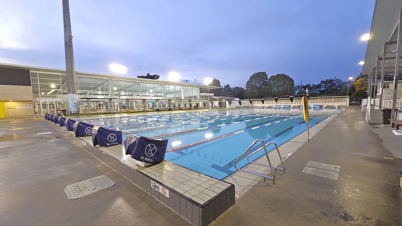In the middle is the 50m 8-lane lap pool equipped with handrails. Diving boards with covers are arranged on the raised pool deck. A free standing banner stands on the pool deck. Floating ropes and blue buntings run across the pool. To the right is an extensive tiered seating with shade. To the left is the facility building with glass walls. Shade structures, seating, and rubbish bins are visible in the grass area. In the middle is the 50m 8-lane lap pool equipped with handrails. Diving boards with covers are arranged on the raised pool deck. A free standing banner stands on the pool deck. Floating ropes and blue buntings run across the pool. To the right is an extensive tiered seating with shade. To the left is the facility building with glass walls. Shade structures, seating, and rubbish bins are visible in the grass area.