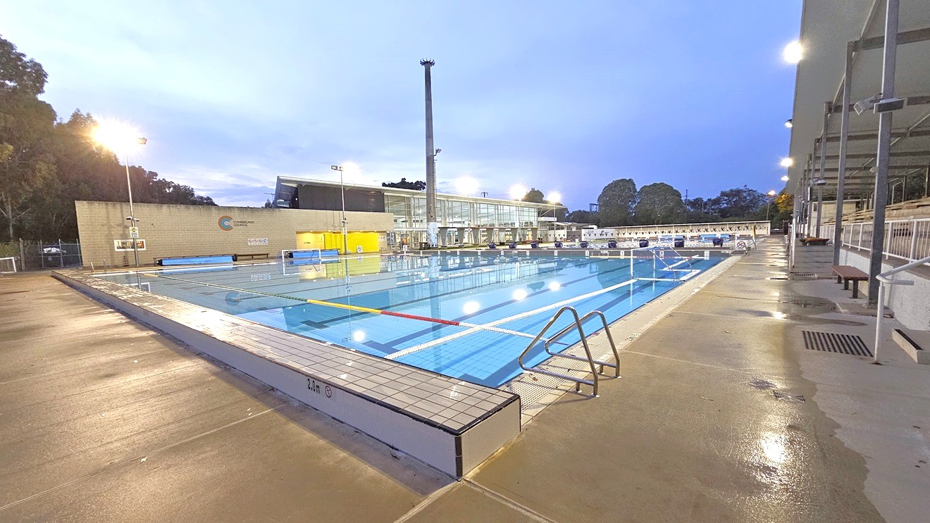 In the middle is a dedicated polo sports pool equipped with handrails. In the middle is the goal set for the polo game. On the far end, diving boards with covers are arranged on the raised pool deck. Floating ropes run across the pool. To the right is an extensive tiered seating with shade. To the far left is the facility building with glass and brick walls. Tall floodlights are also visible. In the middle is a dedicated polo sports pool equipped with handrails. In the middle is the goal set for the polo game. On the far end, diving boards with covers are arranged on the raised pool deck. Floating ropes run across the pool. To the right is an extensive tiered seating with shade. To the far left is the facility building with glass and brick walls. Tall floodlights are also visible.