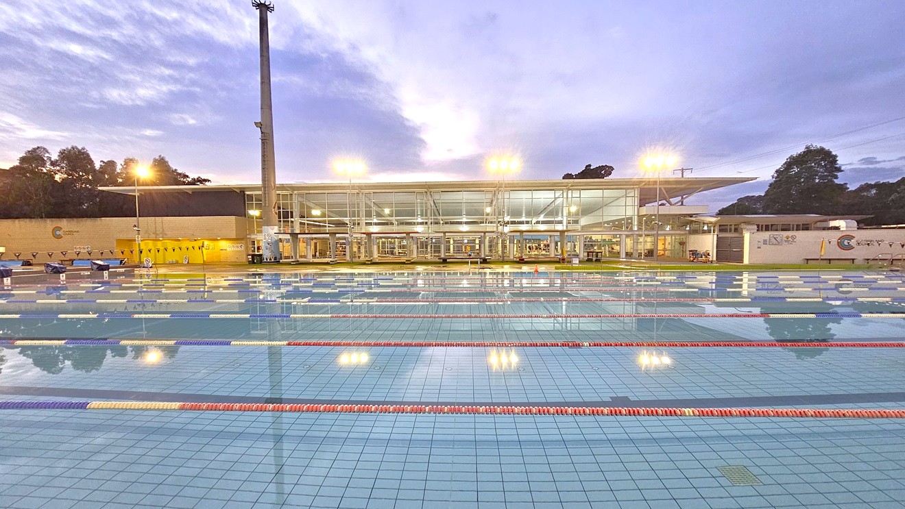 In the middle is the 50m 8-lane lap pool. Floating ropes and blue buntings run across the pool. Diving boards with covers are aligned on the raised pool deck on the left. Beyond the pool is the facility building featuring glass and brick walls. Shade structures are positioned in the manicured grass area in front of the building. Tall floodlights are positioned near the pool. In the middle is the 50m 8-lane lap pool. Floating ropes and blue buntings run across the pool. Diving boards with covers are aligned on the raised pool deck on the left. Beyond the pool is the facility building featuring glass and brick walls. Shade structures are positioned in the manicured grass area in front of the building. Tall floodlights are positioned near the pool.