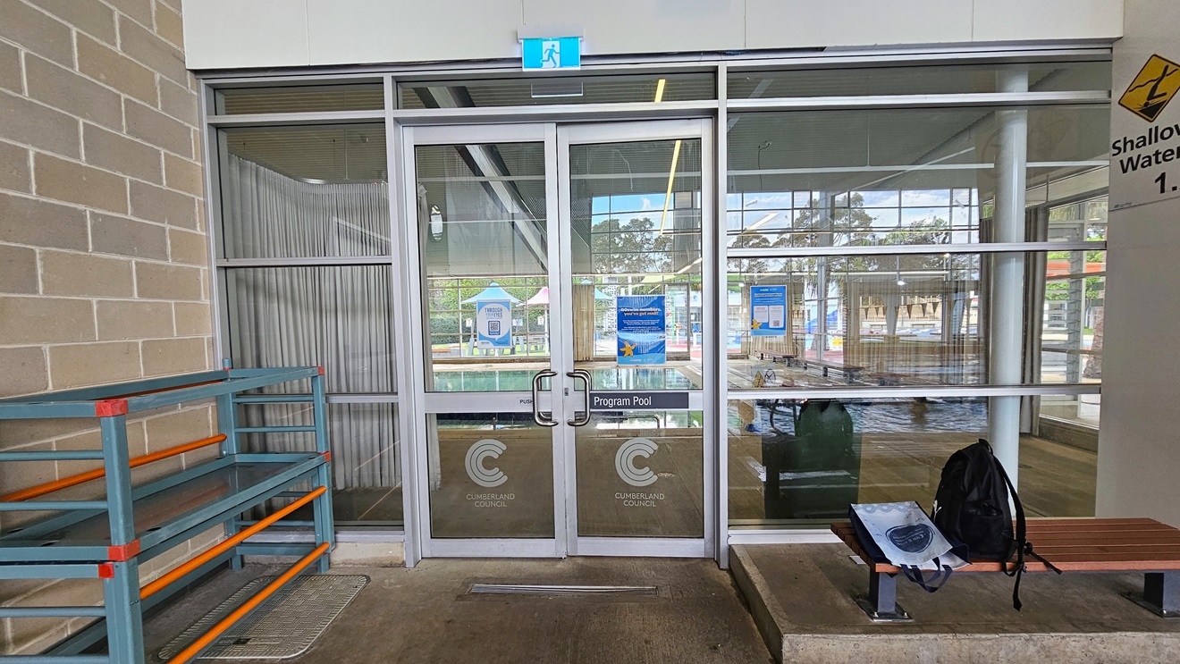 Entrance to the Program Pool is through an outward-opening glass door with hand grabs and facility stickers. A fire exit sign is mounted on top. To the left is a storage rack for belongings. To the right is a bench chair, positioned on a slightly elevated concrete platform against the glass wall. A backpack is placed on top. The upper right wall shows a "Shallow water" sign, and the left wall has brown bricks. Entrance to the Program Pool is through an outward-opening glass door with hand grabs and facility stickers. A fire exit sign is mounted on top. To the left is a storage rack for belongings. To the right is a bench chair, positioned on a slightly elevated concrete platform against the glass wall. A backpack is placed on top. The upper right wall shows a "Shallow water" sign, and the left wall has brown bricks.