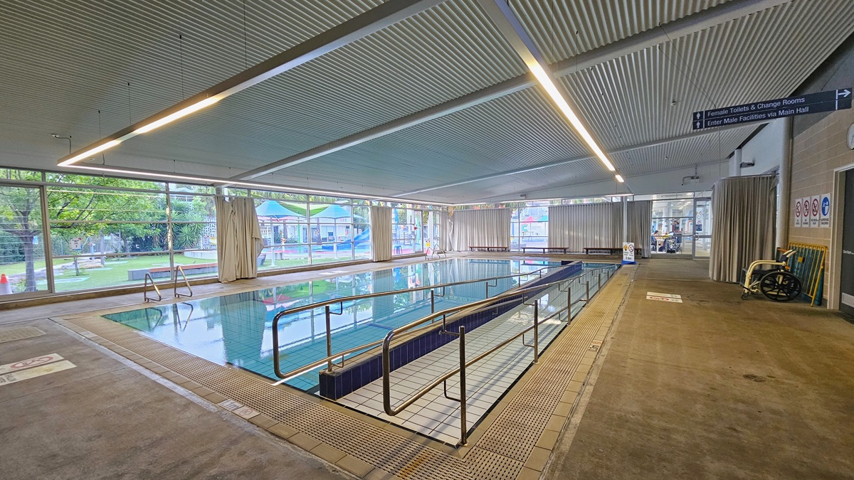 The Program Pool has a ramp entry with handrails on the right. A freestanding banner is positioned in the corner, and overflow grids line the pool. The left wall displays brown bricks and posters, while the right wall features glass with curtains. An outward-opening glass door leads to the Splash Pool. An aquatic wheelchair is placed near the brick wall on the right. The low-level corrugated ceiling is equipped with fluorescent lights, forming a continuous strip above the pool's perimeter. The Program Pool has a ramp entry with handrails on the right. A freestanding banner is positioned in the corner, and overflow grids line the pool. The left wall displays brown bricks and posters, while the right wall features glass with curtains. An outward-opening glass door leads to the Splash Pool. An aquatic wheelchair is placed near the brick wall on the right. The low-level corrugated ceiling is equipped with fluorescent lights, forming a continuous strip above the pool's perimeter.