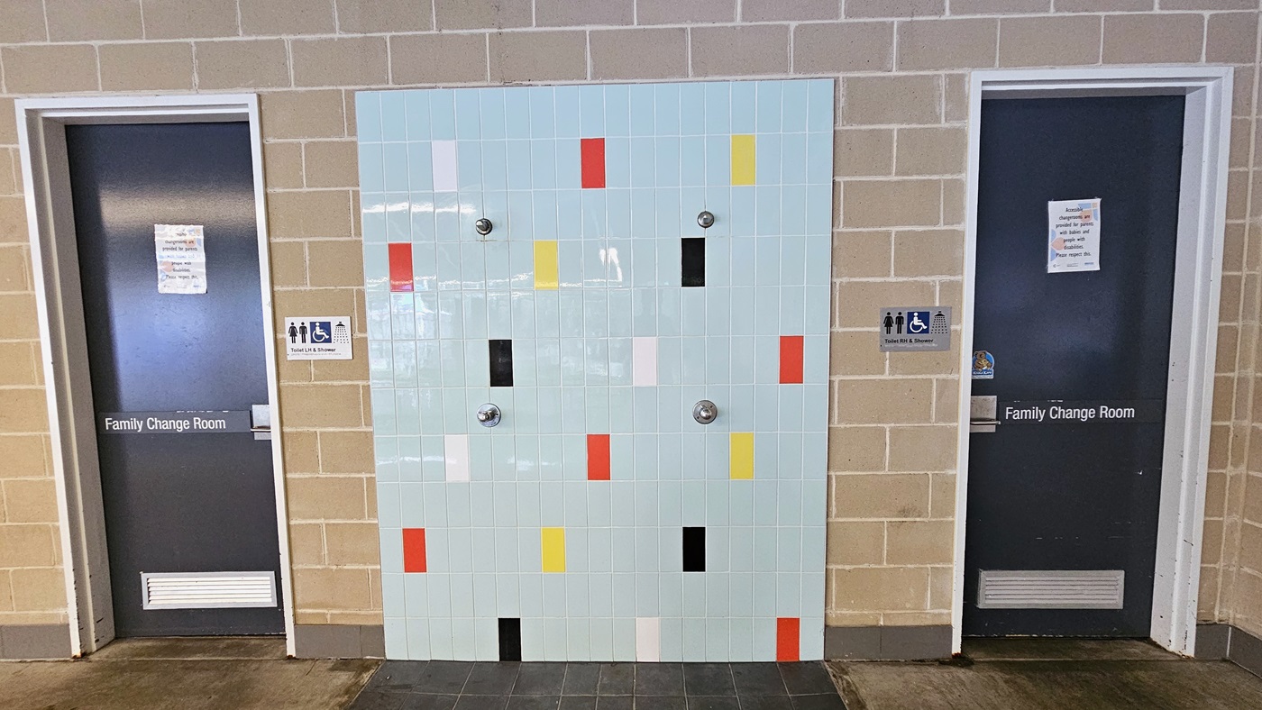 In the center of a brown brick wall is an outdoor shower with a wall adorned with multi-color tiles. Two grey doors, each marked with a "Family Change Room" sign, are present. Additional signage indicating accessibility, showers, and all-gender access is also mounted on the walls. In the center of a brown brick wall is an outdoor shower with a wall adorned with multi-color tiles. Two grey doors, each marked with a "Family Change Room" sign, are present. Additional signage indicating accessibility, showers, and all-gender access is also mounted on the walls.