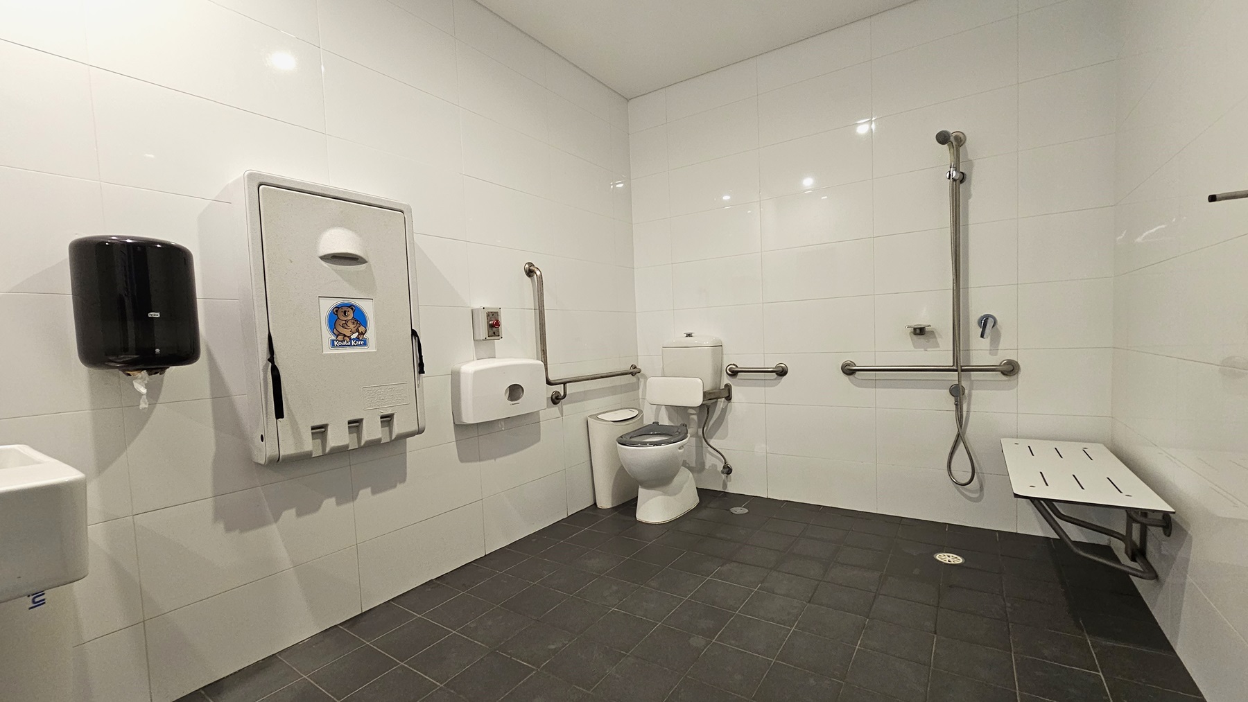 Inside the accessible change room, on the left is a toilet bowl equipped with hand grabs, accompanied by a rubbish bin. A tissue dispenser, a foldable change table, and a liquid hand soap dispenser are affixed to the left wall. A washbasin and a poster are mounted on the wall. To the right is a shower with handrails, and a bench is positioned against the wall. The floor is covered with grey tiles, and the walls and ceiling are white. Inside the accessible change room, on the left is a toilet bowl equipped with hand grabs, accompanied by a rubbish bin. A tissue dispenser, a foldable change table, and a liquid hand soap dispenser are affixed to the left wall. A washbasin and a poster are mounted on the wall. To the right is a shower with handrails, and a bench is positioned against the wall. The floor is covered with grey tiles, and the walls and ceiling are white.