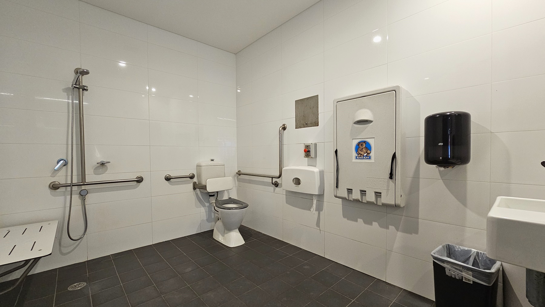 Inside the accessible change room, on the right is a toilet bowl equipped with hand grabs, accompanied by a rubbish bin. A tissue dispenser, a foldable change table, and a liquid hand soap dispenser are affixed to the right wall. A washbasin and a poster are mounted on the wall. To the left is a shower with handrails, and a bench is positioned against the wall. The floor is covered with grey tiles, and the walls and ceiling are white. Inside the accessible change room, on the right is a toilet bowl equipped with hand grabs, accompanied by a rubbish bin. A tissue dispenser, a foldable change table, and a liquid hand soap dispenser are affixed to the right wall. A washbasin and a poster are mounted on the wall. To the left is a shower with handrails, and a bench is positioned against the wall. The floor is covered with grey tiles, and the walls and ceiling are white.