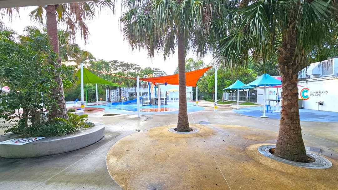 The outdoor Splash Park features palm trees and bright orange and green shade cloths. In the center is a blue slide with tipping buckets, water sprays, geysers, waterfalls, and fountains. Plastic tables and chairs are arranged under large blue umbrella shades on the right. The floor offers different shapes and colors. On the left, plants and trees are inside a round concrete planter box along with a safety sign. Further ahead, trees are visible. The outdoor Splash Park features palm trees and bright orange and green shade cloths. In the center is a blue slide with tipping buckets, water sprays, geysers, waterfalls, and fountains. Plastic tables and chairs are arranged under large blue umbrella shades on the right. The floor offers different shapes and colors. On the left, plants and trees are inside a round concrete planter box along with a safety sign. Further ahead, trees are visible.