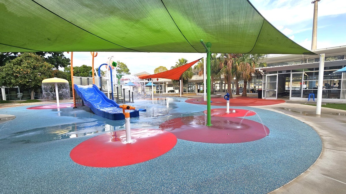 The Splash Park includes a blue slide, tipping buckets, water sprays, geysers, waterfalls, and fountains. Green and orange shade cloths offer shading. Tall palm and other trees border the left and right edges. The ground displays diverse shapes and colors. In the distance, a white building with a glass wall is visible on the right. The Splash Park includes a blue slide, tipping buckets, water sprays, geysers, waterfalls, and fountains. Green and orange shade cloths offer shading. Tall palm and other trees border the left and right edges. The ground displays diverse shapes and colors. In the distance, a white building with a glass wall is visible on the right.