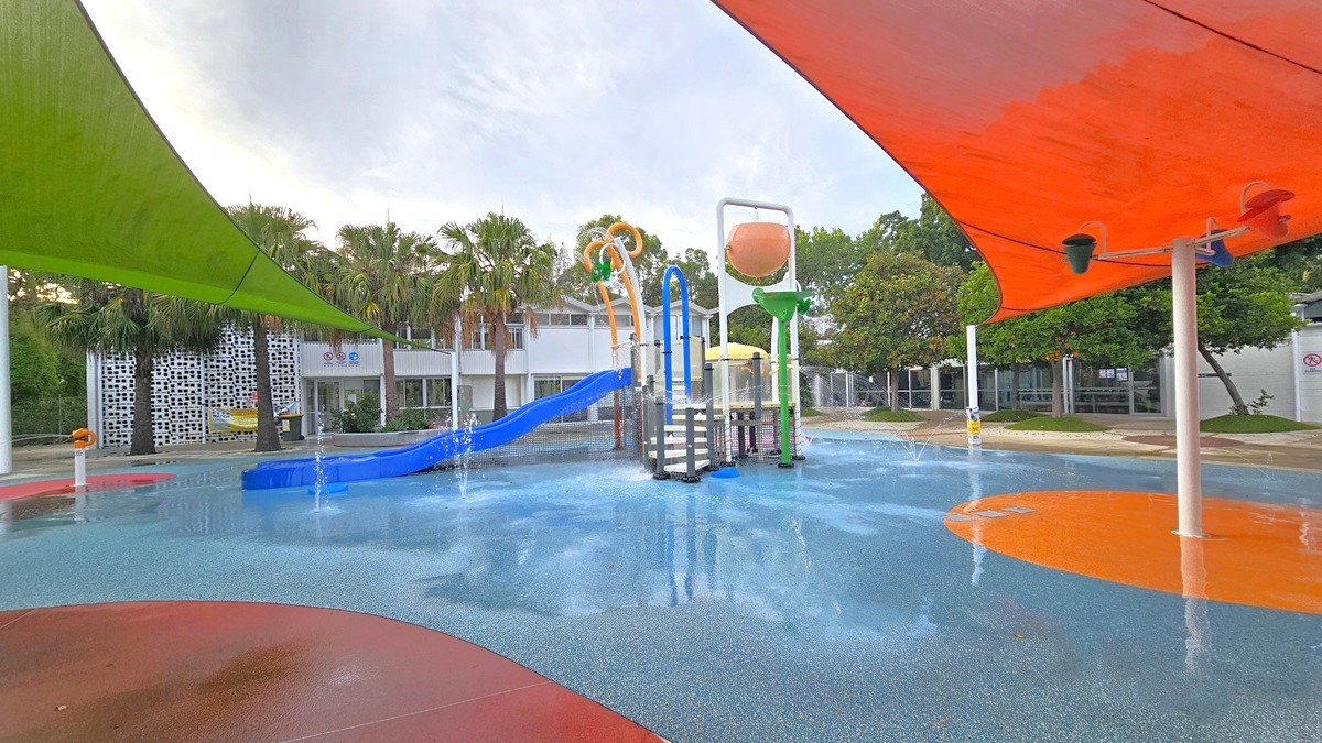 The Splash Park includes a blue slide, tipping buckets, water sprays, geysers, waterfalls, and fountains. Green and orange shade cloths provide shading. Tall palms and other trees border the left edges. The ground exhibits diverse shapes and colors. In the background, there's a white building with a glass wall. To the right, trees and neatly trimmed grass are visible. The Splash Park includes a blue slide, tipping buckets, water sprays, geysers, waterfalls, and fountains. Green and orange shade cloths provide shading. Tall palms and other trees border the left edges. The ground exhibits diverse shapes and colors. In the background, there's a white building with a glass wall. To the right, trees and neatly trimmed grass are visible.