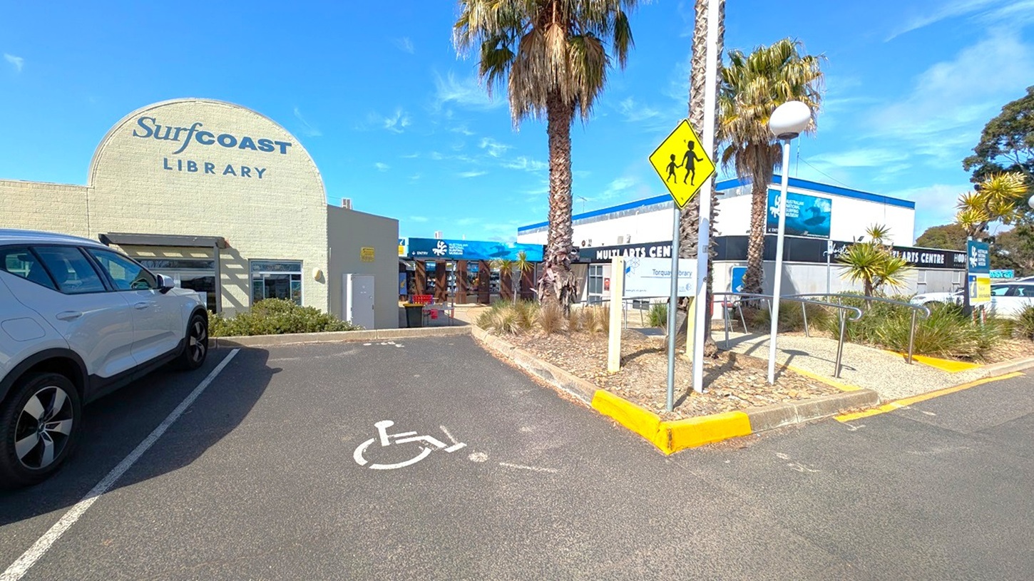 A car park with a white car on the right. In the centre, there are palm trees and a pedestrian crossing sign. A sign for visitor information is visible near the trees. On the left, several parked cars are lined up. In the background, a building with signage for a library and a blue storefront can be seen. The ground is paved, and there are designated parking spaces, including one for disabled parking.