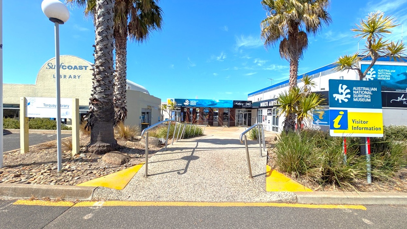 Pathway leading to a building with signs for the Australian National Surfing Museum and Visitor Information on the right. On the left, there's a sign for the Surf Coast Library. Palm trees and shrubs line the path, with metal railings on either side. The ground is a mix of pebbles and concrete, and the buildings have a modern design with large windows.