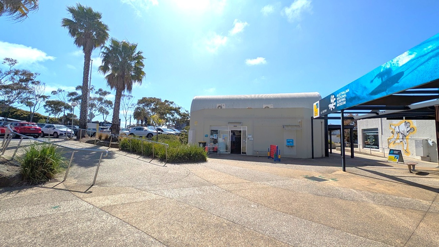 A paved area with a building in the centre, featuring a curved roof and an open entrance. To the right, a sign reads "Australian National Surfing Museum" with a surfing image. A mural of a surfer is painted on the wall. On the left, there are palm trees and parked cars. The ground is a mix of textured concrete and green patches, with metal railings and some low bushes.