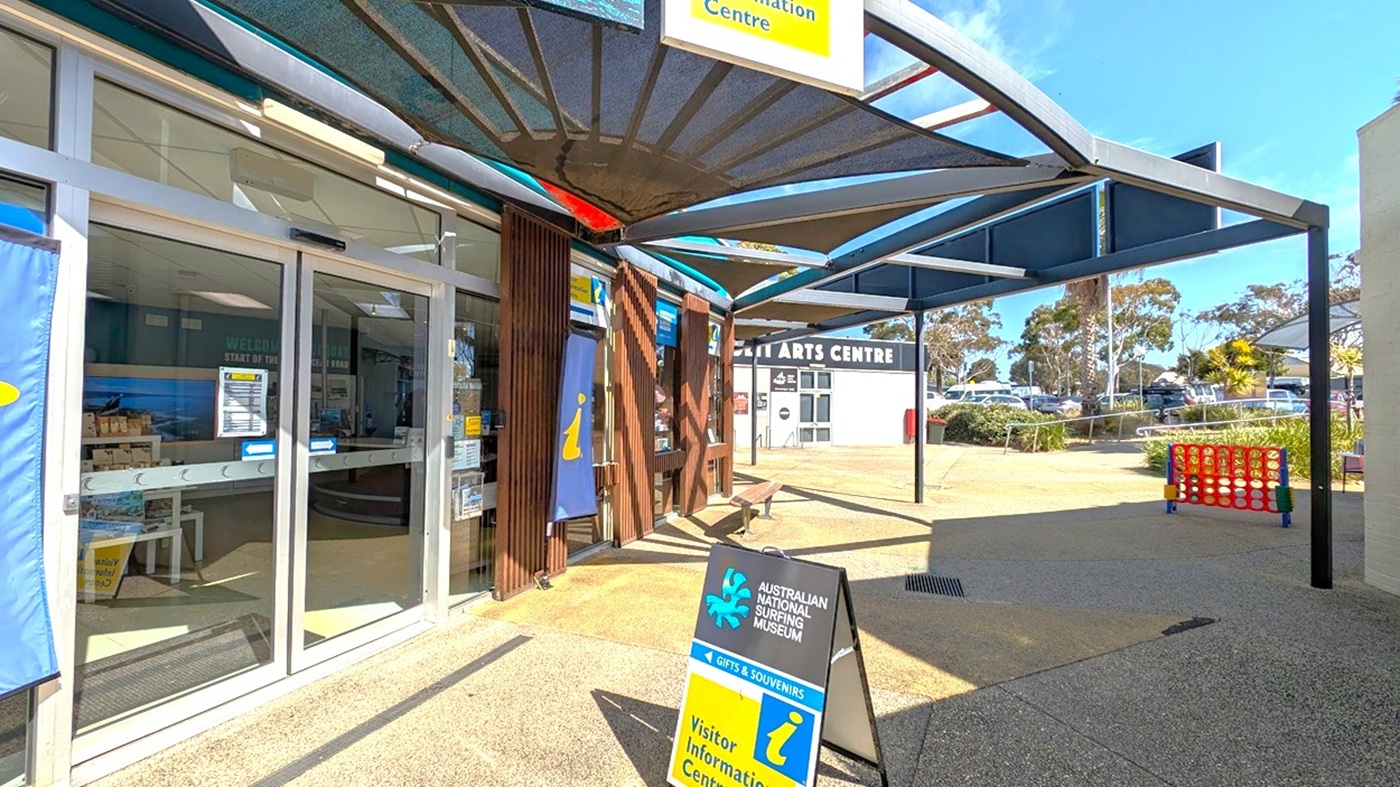 Entrance to the Australian National Surfing Museum on the left, with glass doors and informational banners. A signboard in front reads "Visitor Information Centre." A shaded area with a modern canopy extends overhead. In the background, the Surf City Arts Centre is visible, along with parked cars and trees. The ground is paved, and a large Connect Four game is set up on the right.