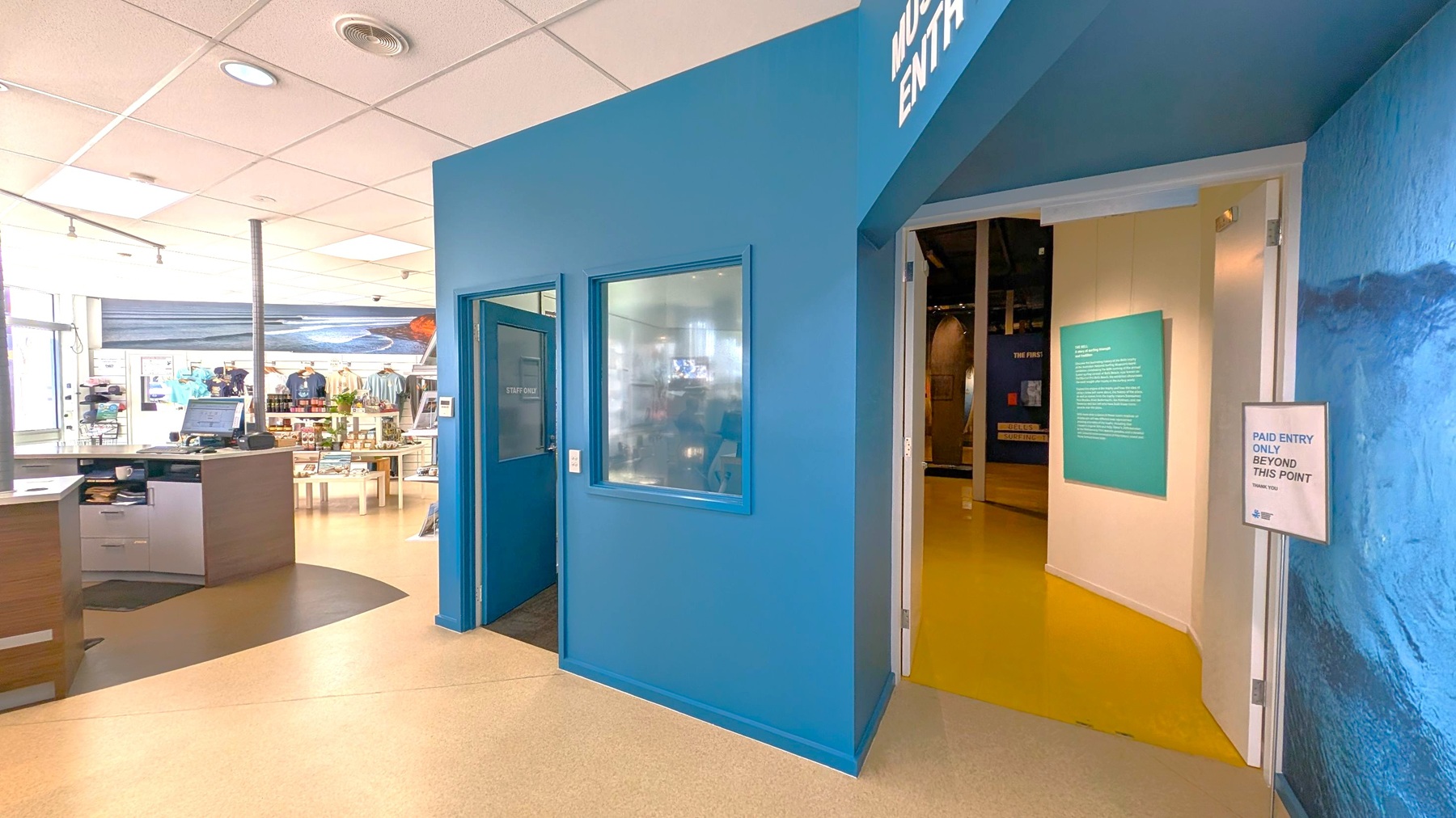 A room with a blue wall on the right, featuring a "Museum Entry" sign above an open door. A sign next to the door reads "Paid Entry Only Beyond This Point." Inside, a yellow floor leads to an exhibit area. On the left, a reception desk with a computer is visible, and beyond it, a shop area with clothing and merchandise on display. The ceiling is white with recessed lighting.